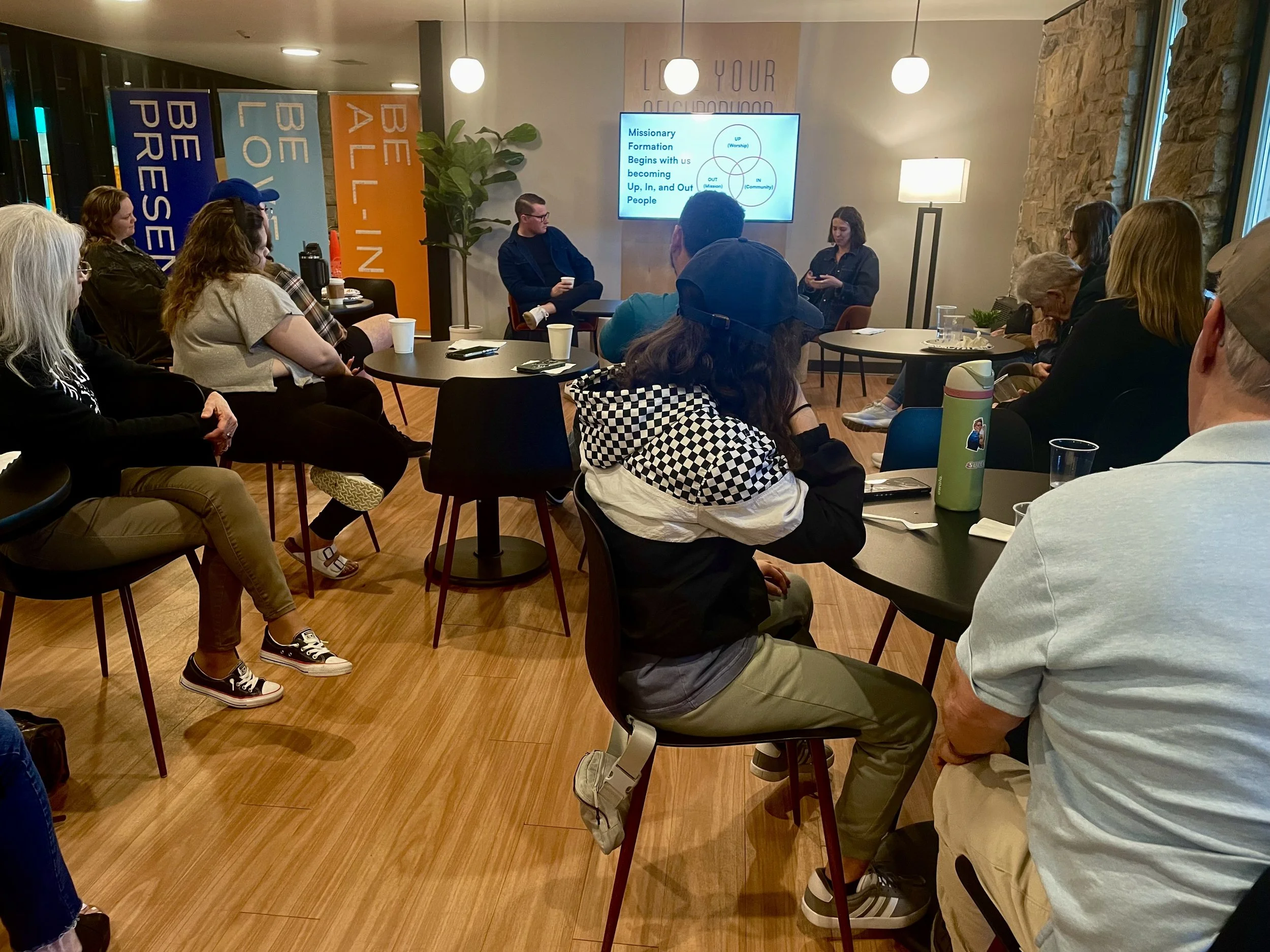 A group of people seated around tables in a room, attending a presentation with a woman speaker and a man presenter, in a room with banners, plants, a stone wall, and a screen displaying a presentation slide.