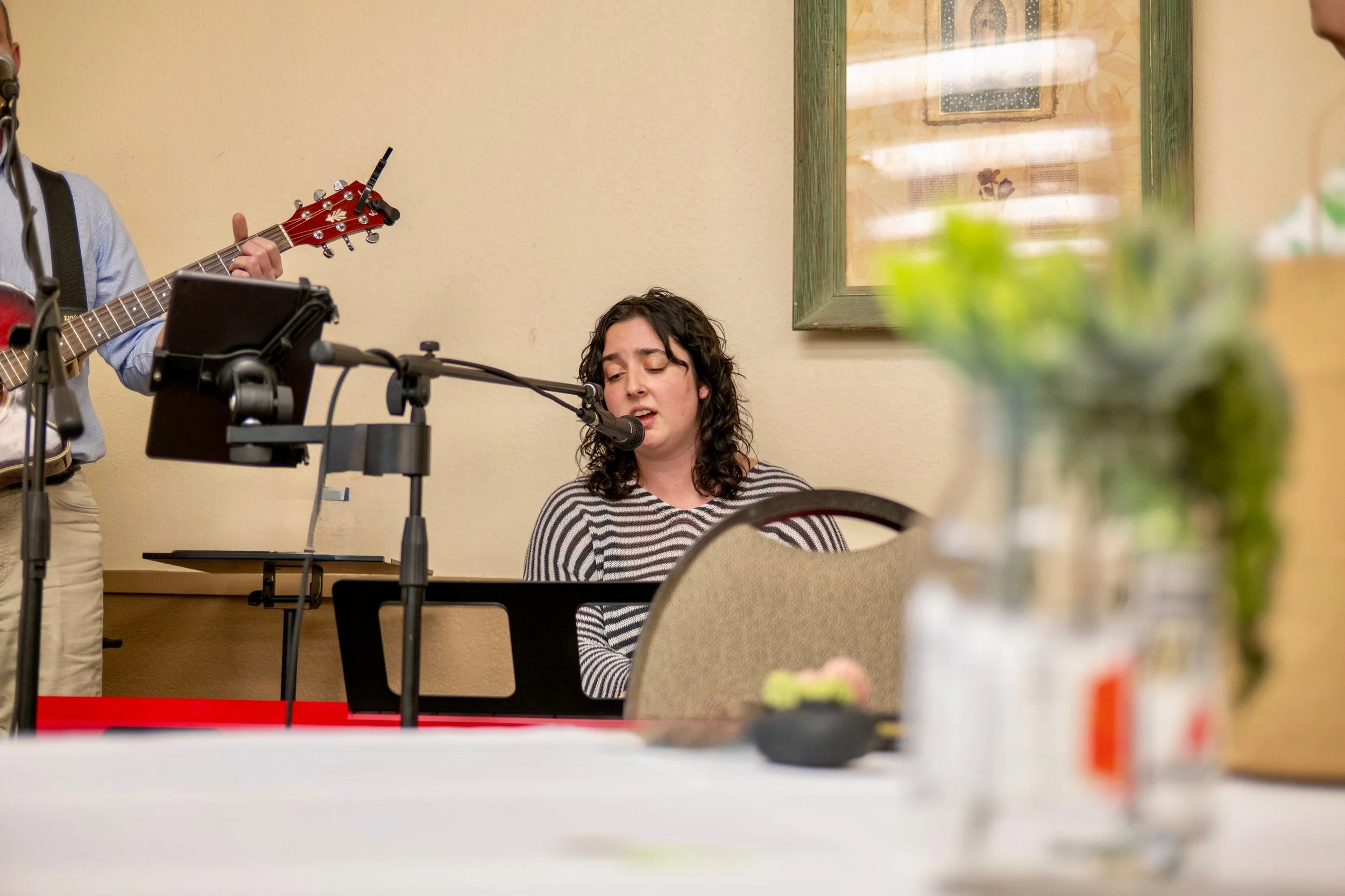 Female singer with curly hair sings into a microphone while musicians play guitar in the background, at an indoor event with a beige wall and framed artwork.