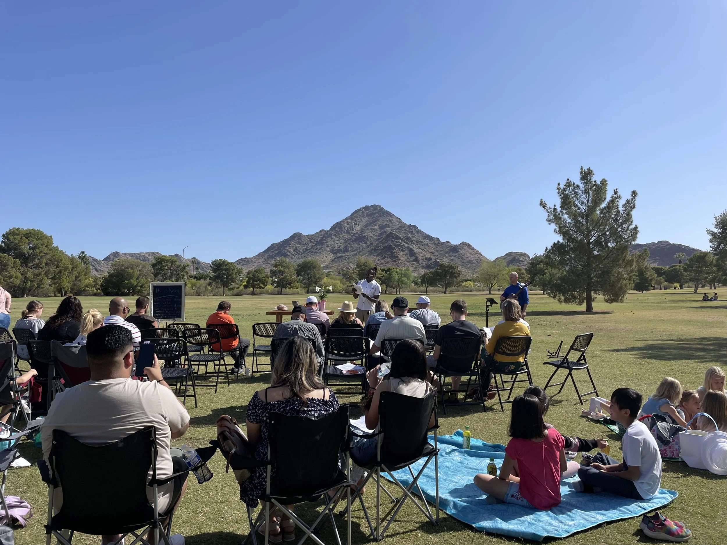 Outdoor gathering with people seated on chairs and on a picnic blanket on grass, with two speakers standing in front of a scenic mountain backdrop under a clear blue sky.