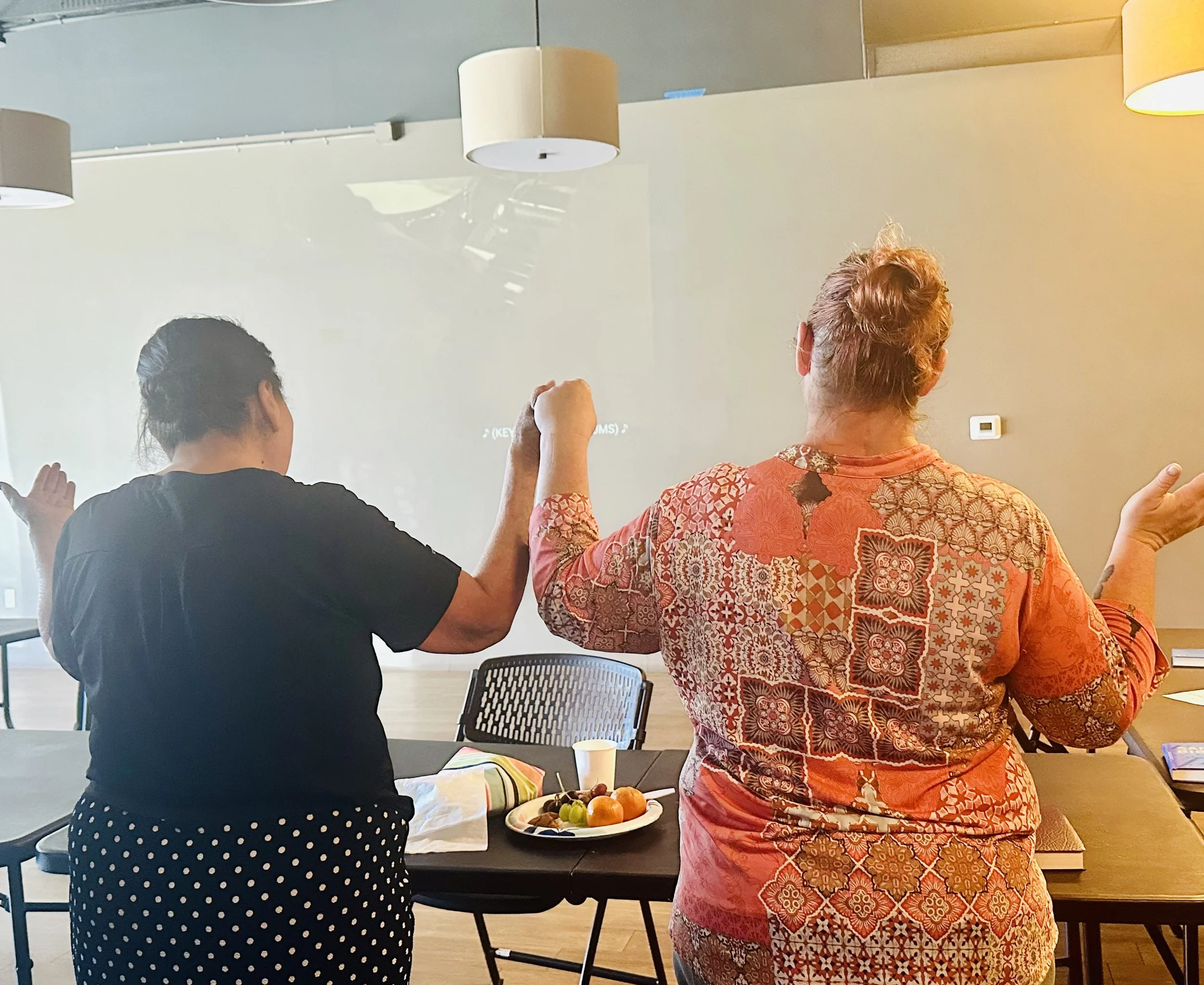 Two women are standing with their backs to the camera, raising their hands in a room with a large projection screen. One woman is wearing a black top and polka dot skirt, and the other has a colorful patterned blouse or dress. A fruit tray and a cup are on the table in front of them.