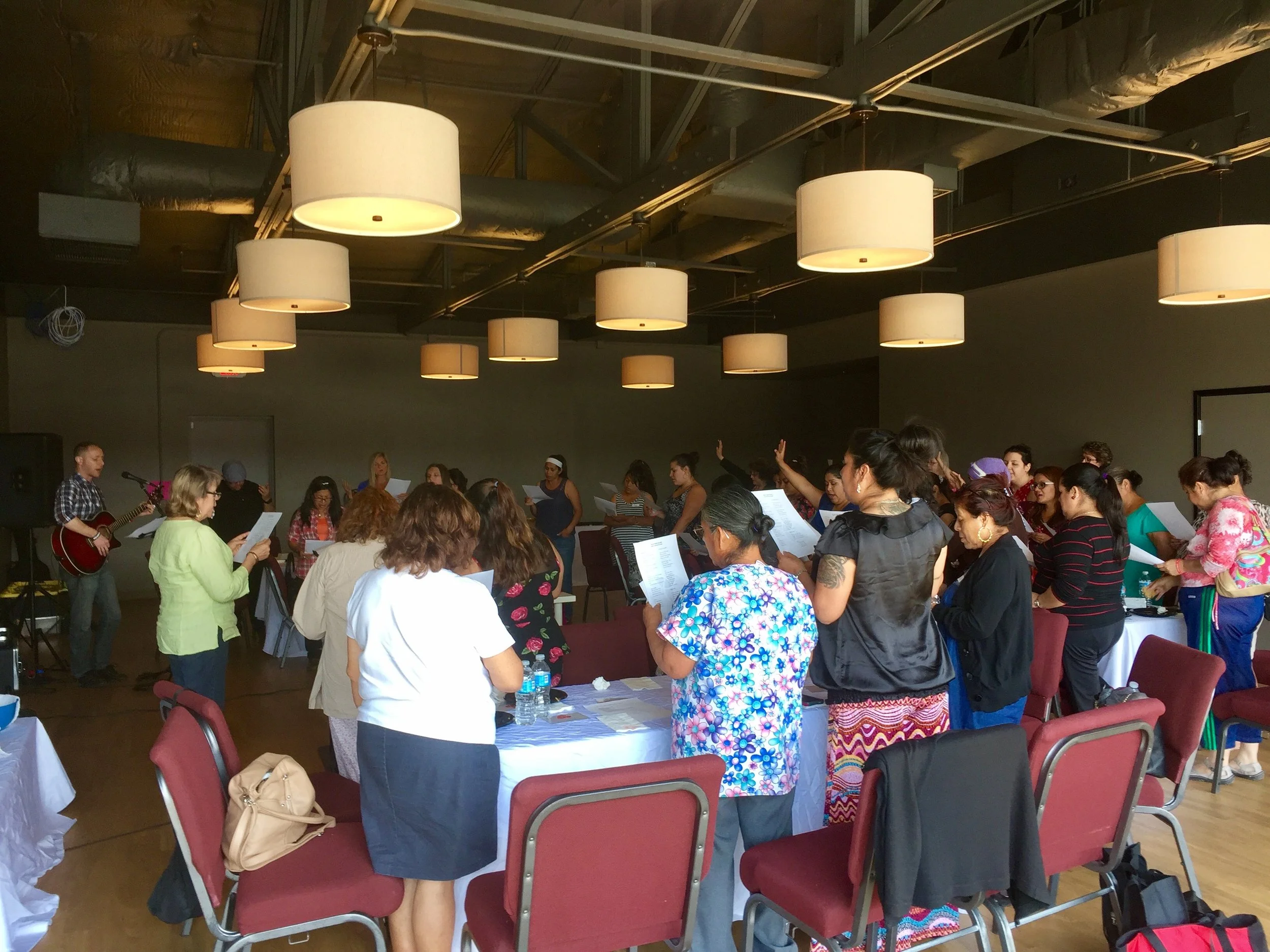 Group of people standing and singing, some holding papers, in a room with hanging lights and a musician playing guitar in the background.