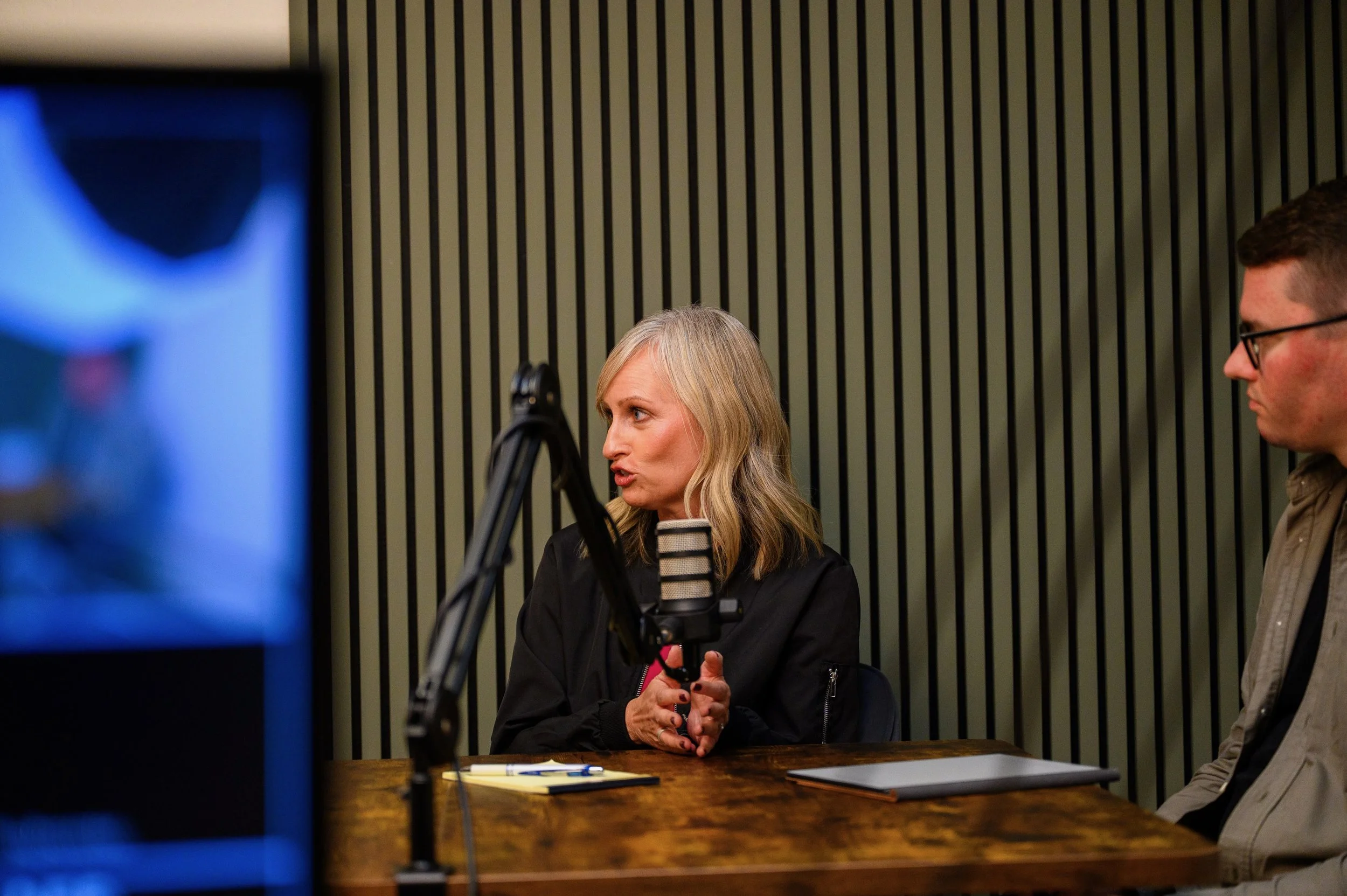 A woman with blonde hair speaking into a microphone during an interview or podcast recording, with a man sitting next to her, in a room with striped wall panels.