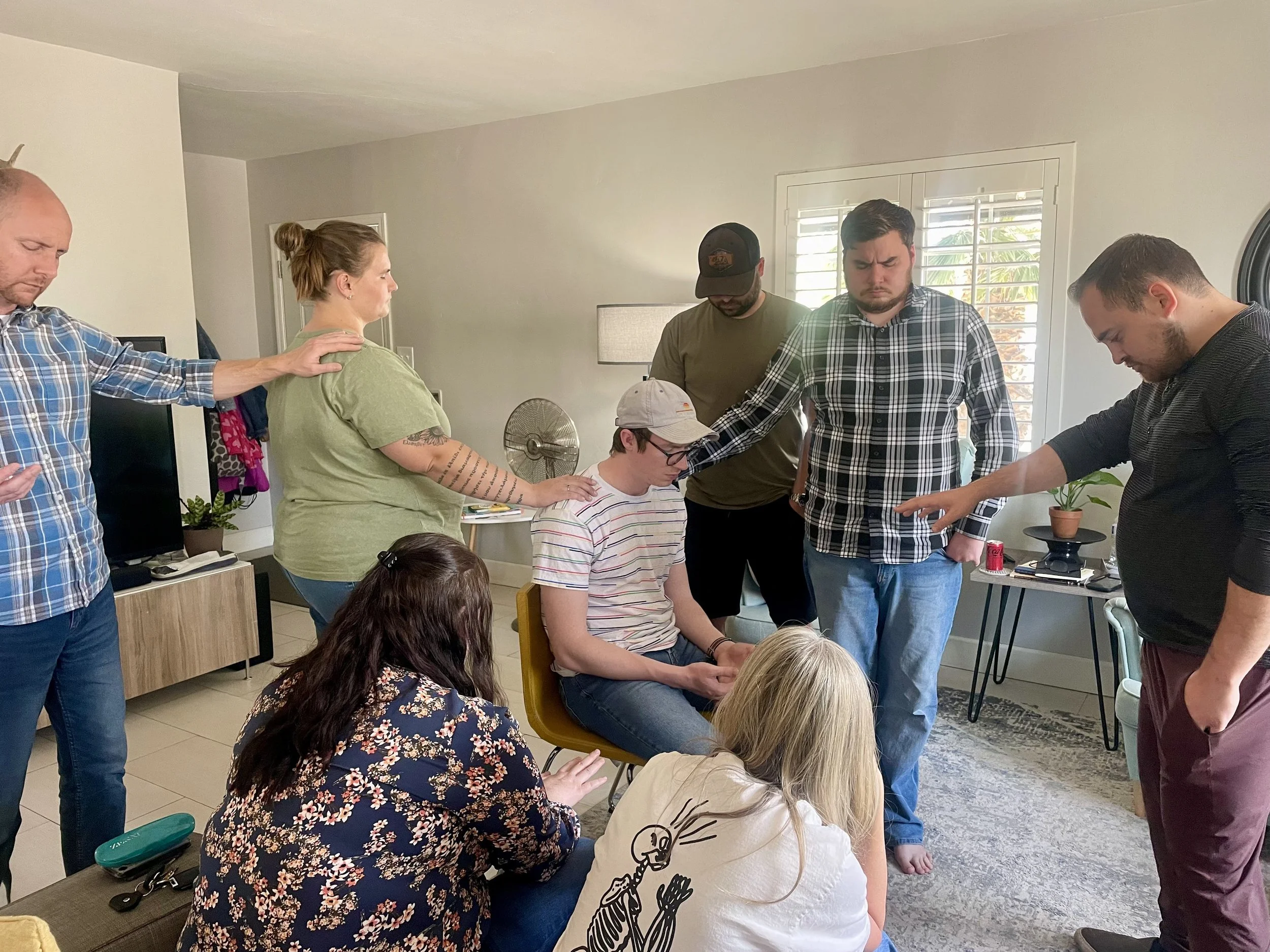 Group of people praying together in a living room, with some standing and some kneeling, eyes closed or looking down, creating a solemn and spiritual atmosphere.