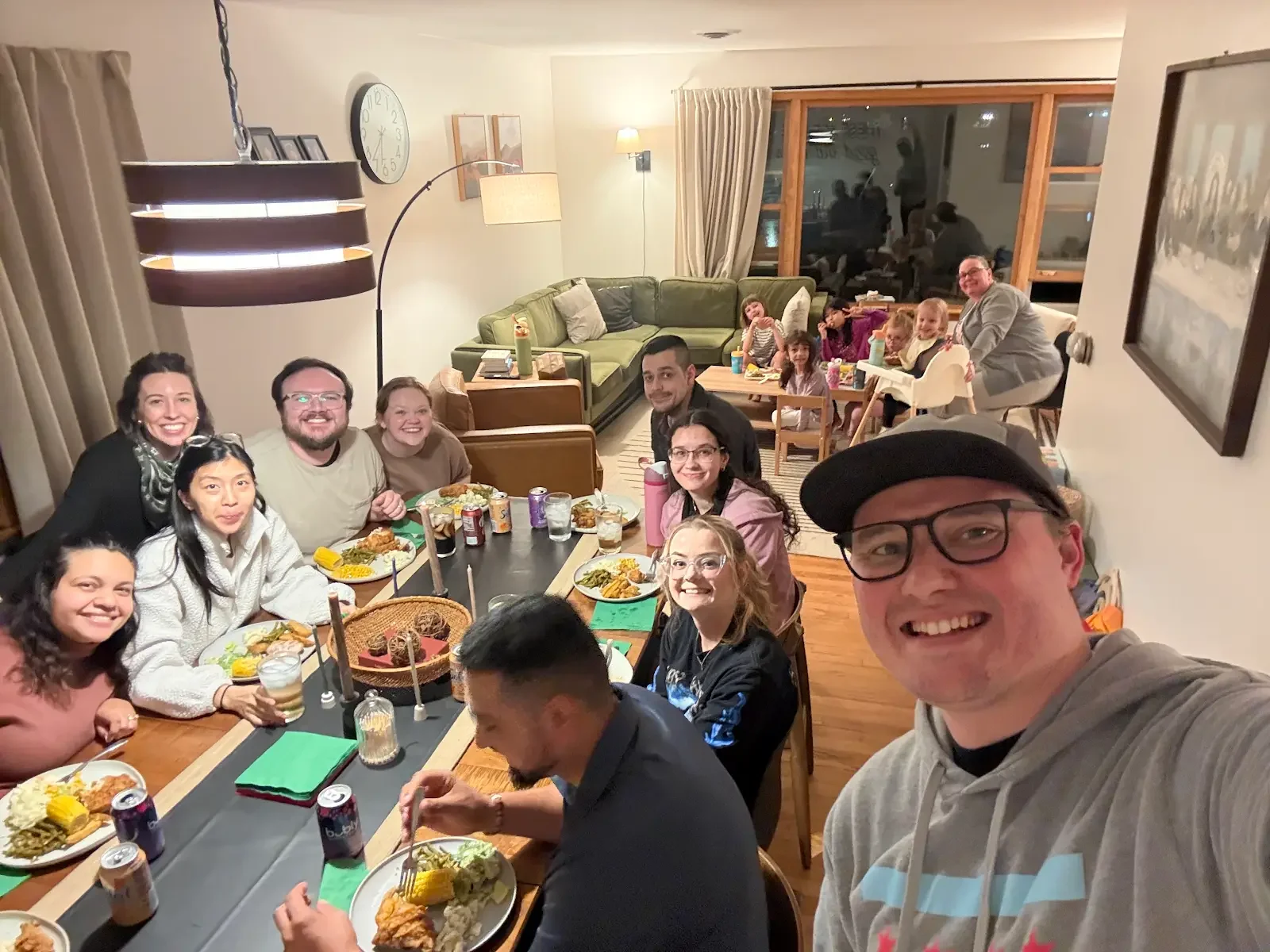 Group of people gathered around a dining table with food, children in high chairs and adults, indoors with a large window and a green couch in the background, celebrating together.
