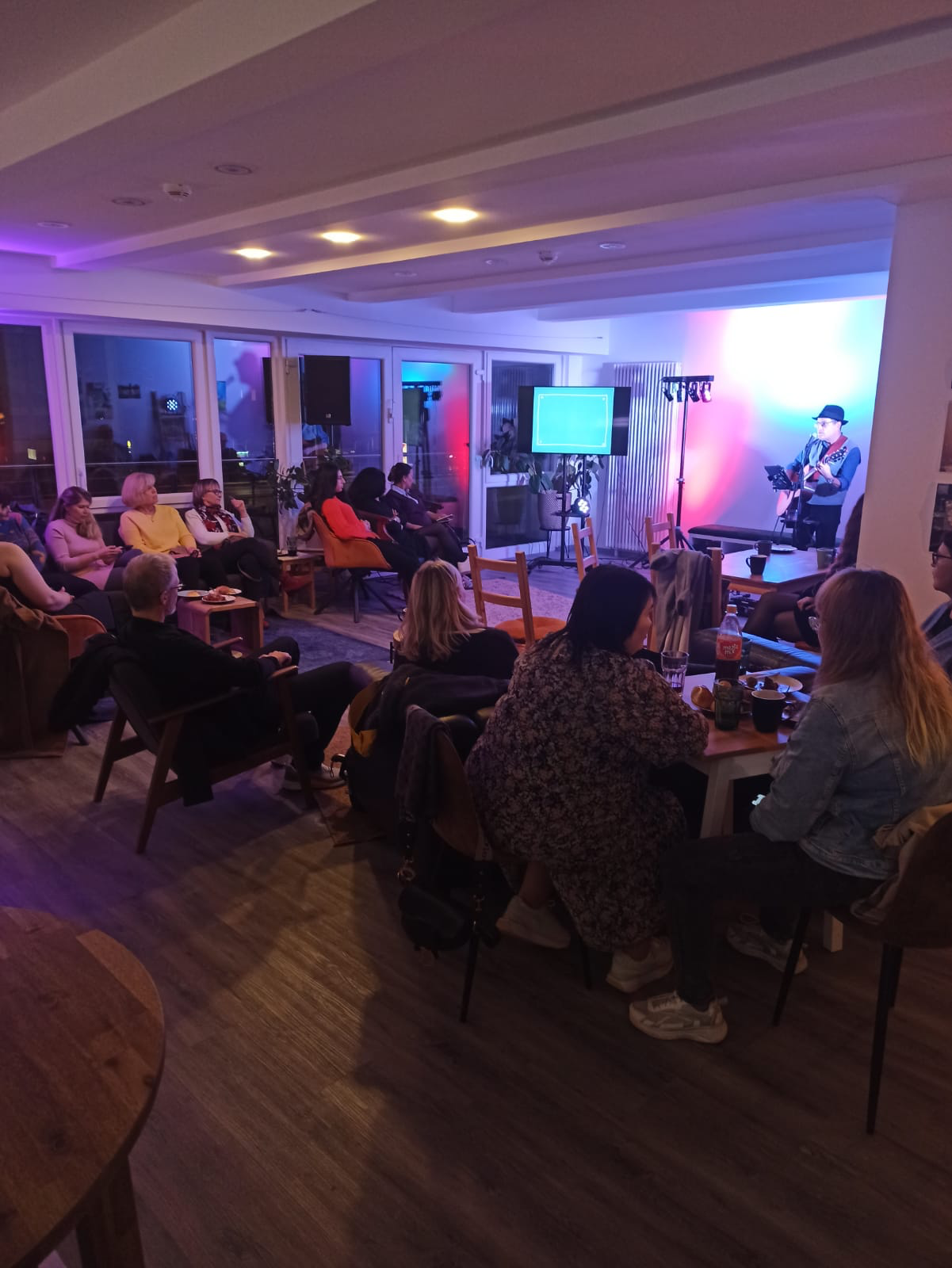 People watching a live music performance in a cozy indoor venue with a singer playing guitar on stage, colorful lighting, and large windows.