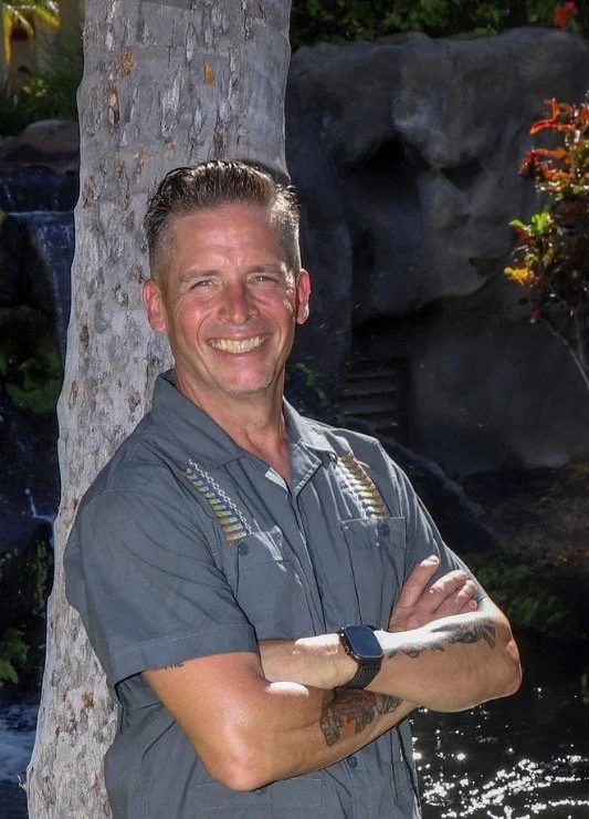 A smiling man standing outdoors next to a tree, crossing his arms, with water and rocks in the background.