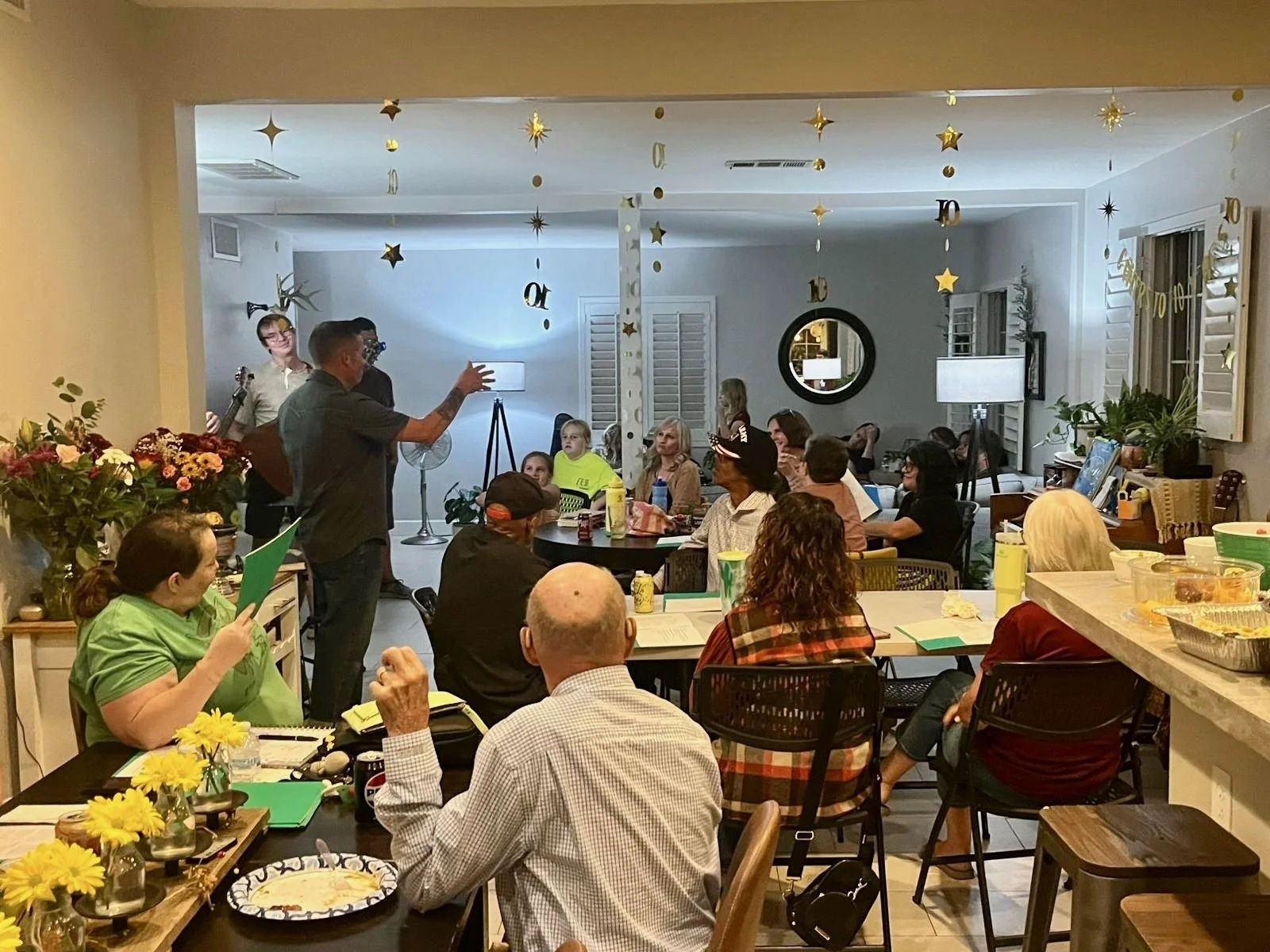 A gathering of people in a decorated living room, with a person speaking to the group, surrounded by festive decorations and a variety of food and drinks.