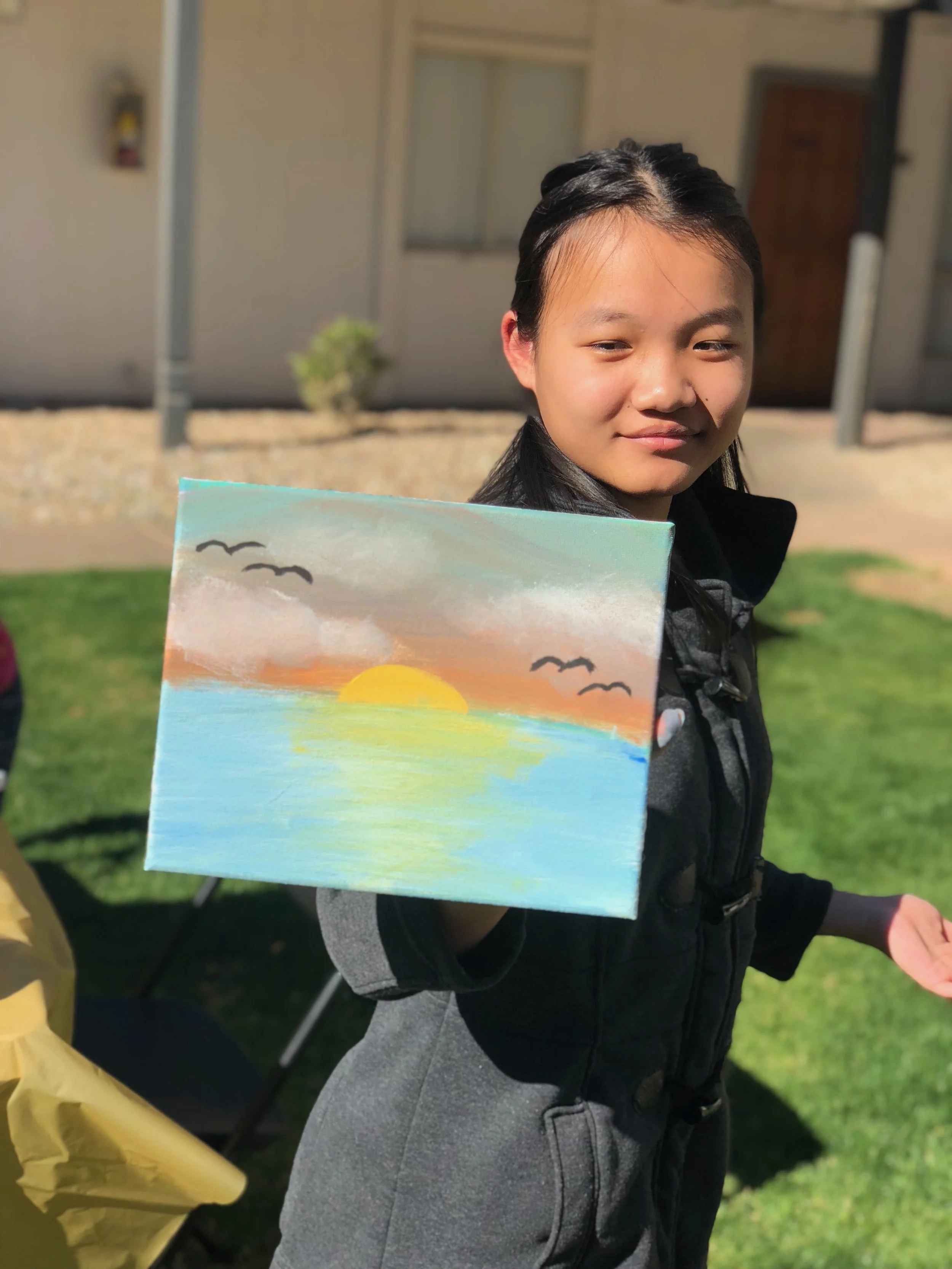 Young girl outdoors holding a painting of a sunset over water with birds flying in the sky.