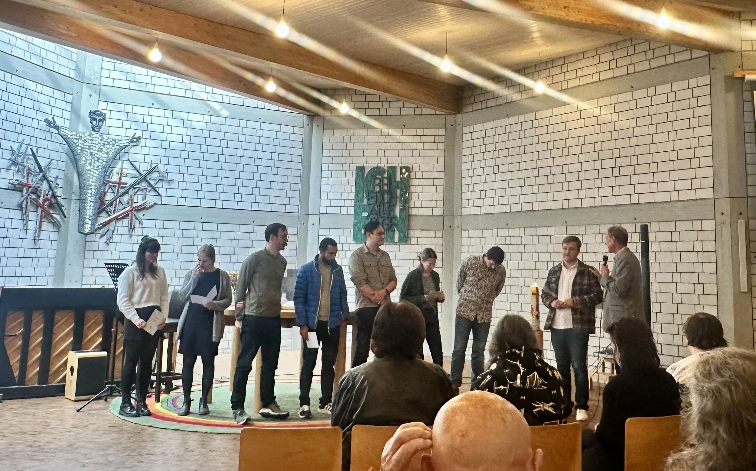 Group of people standing in a semicircle on a stage, participating in a presentation or performance in a room with white brick walls and decorative artwork.