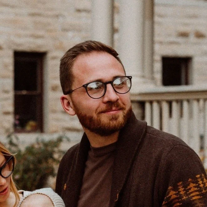 A man with glasses, a beard, and brown hair smiling outdoors with a stone building and balcony in the background.