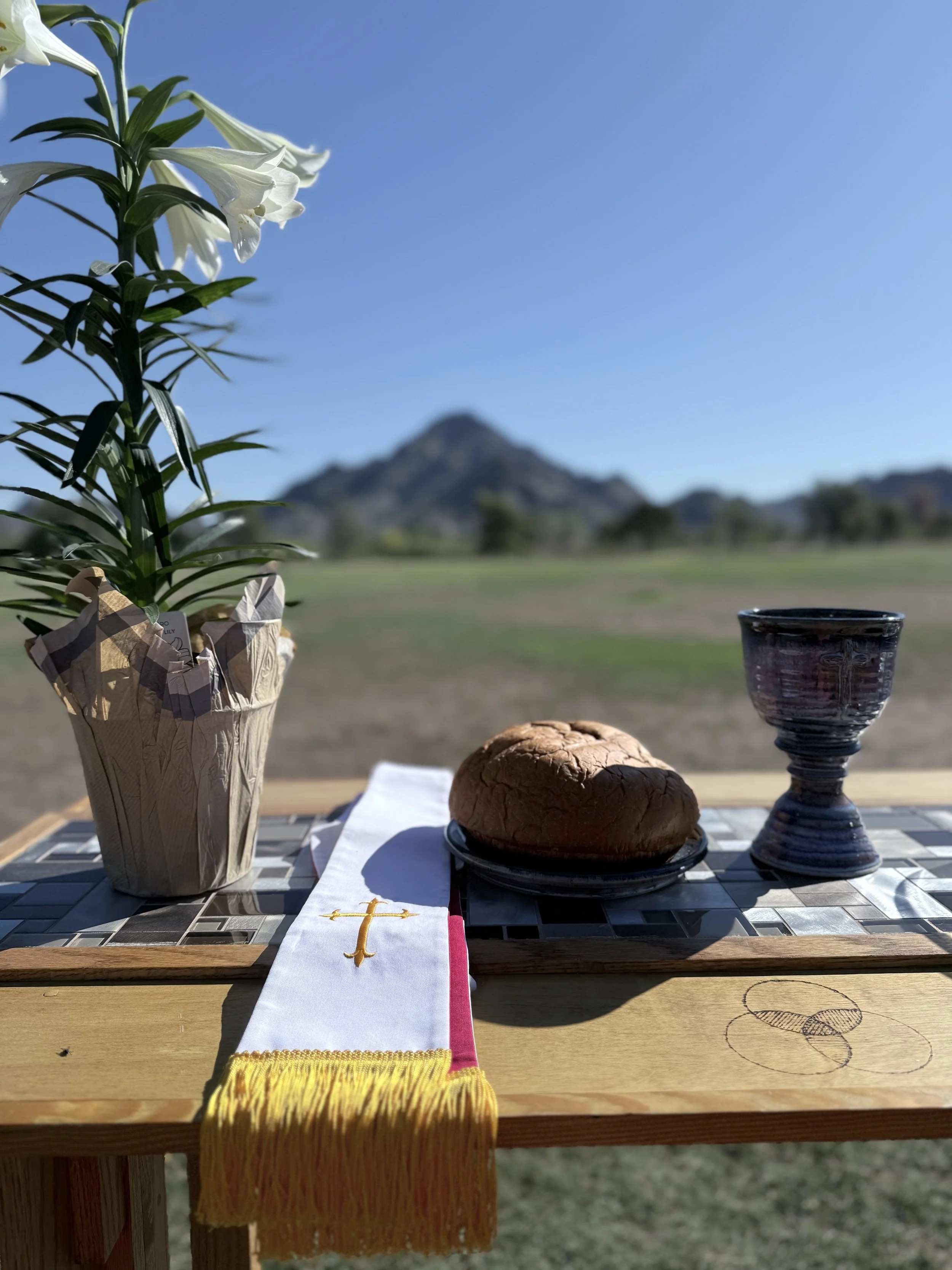 An outdoor table set with a potted white lily flower, a loaf of bread on a plate, a purple ceramic goblet, and a white cloth with a gold cross and yellow fringe, against a backdrop of a clear blue sky and mountain range.
