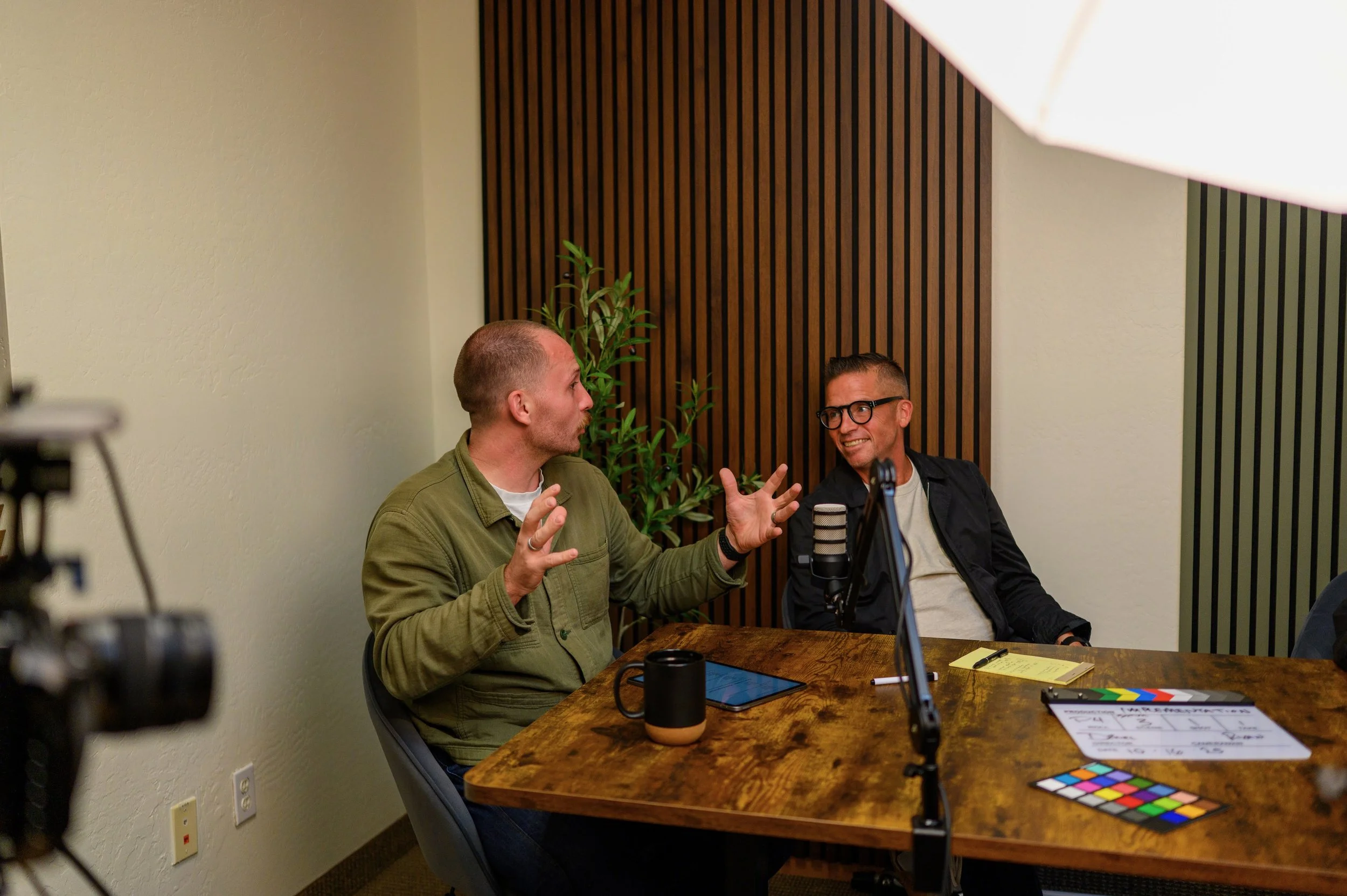 Two men engaged in a discussion during a podcast recording, sitting at a wooden table with recording equipment, notes, and a mug, with a potted plant and wooden wall paneling in the background.