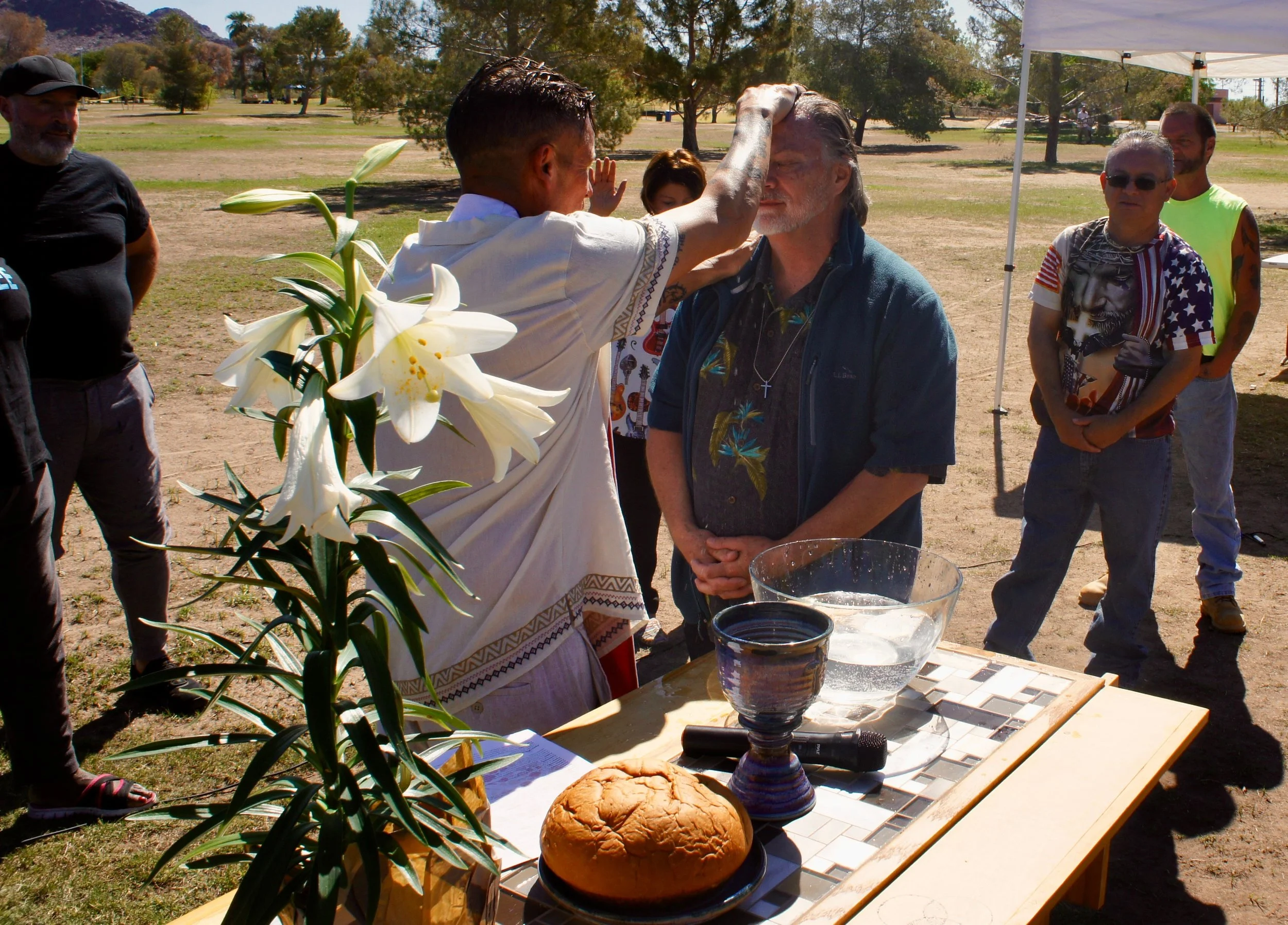 A group of people participating in a baptism ceremony outdoors under a white canopy, with a table holding bread, a water basin, and religious items, surrounded by trees and open field.