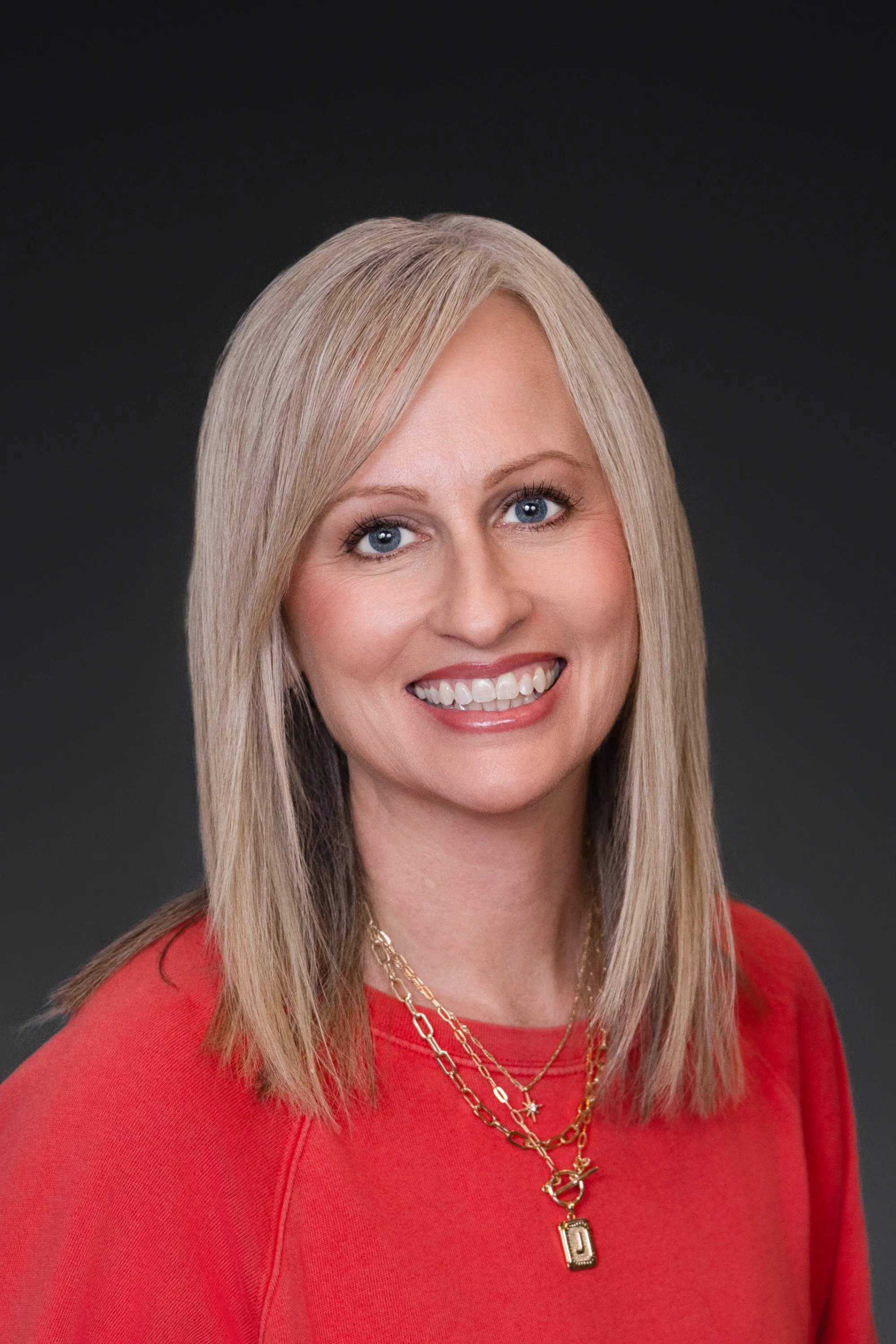 Headshot of a woman with blonde hair, blue eyes, and a bright smile, wearing a red top and layered gold necklaces.