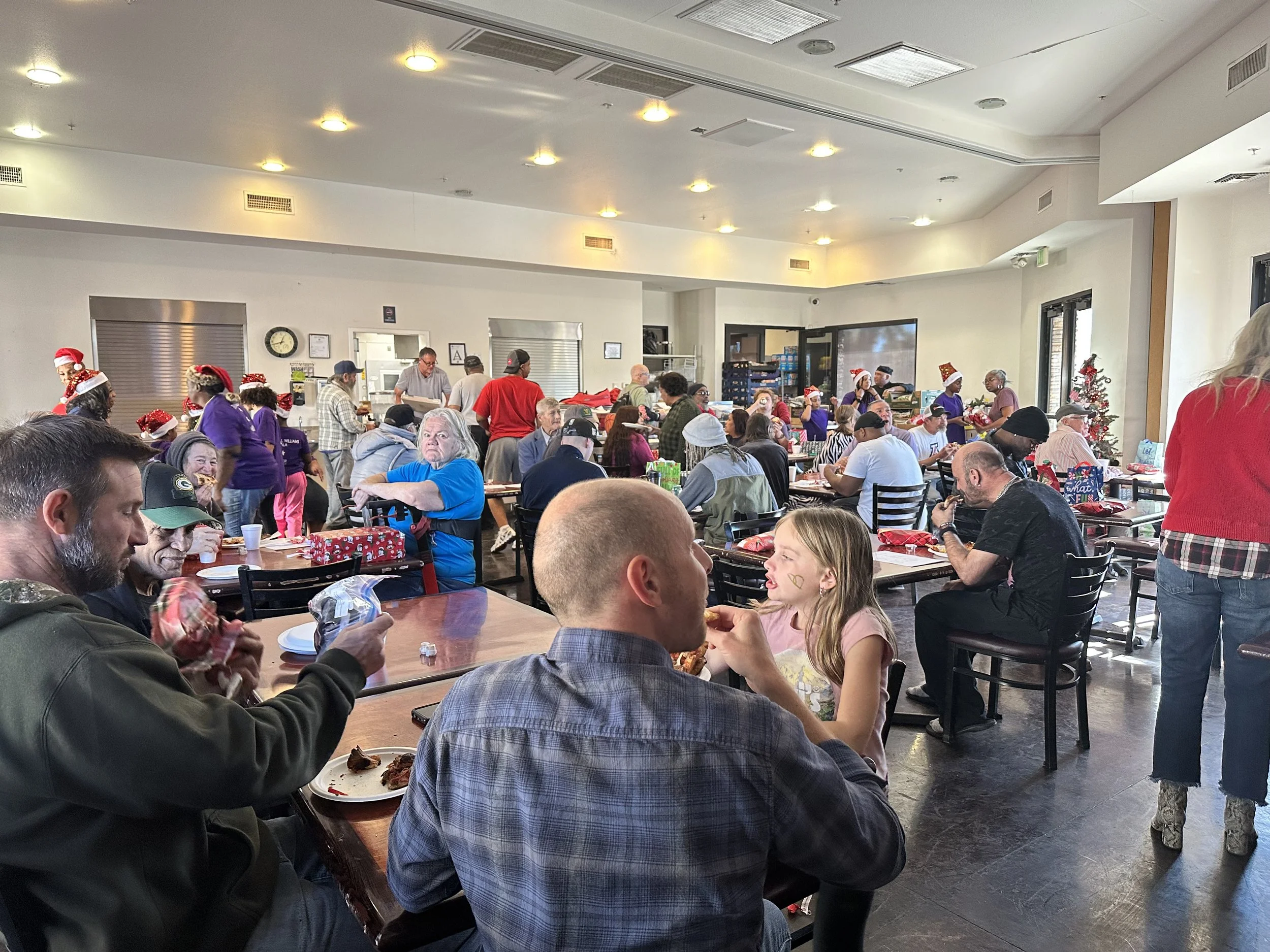 A crowded indoor dining area filled with people sitting at tables, many wearing Christmas hats and decorations, some eating, talking, and socializing. There are holiday decorations, including a Christmas tree, and everyone appears to be celebrating a festive occasion.