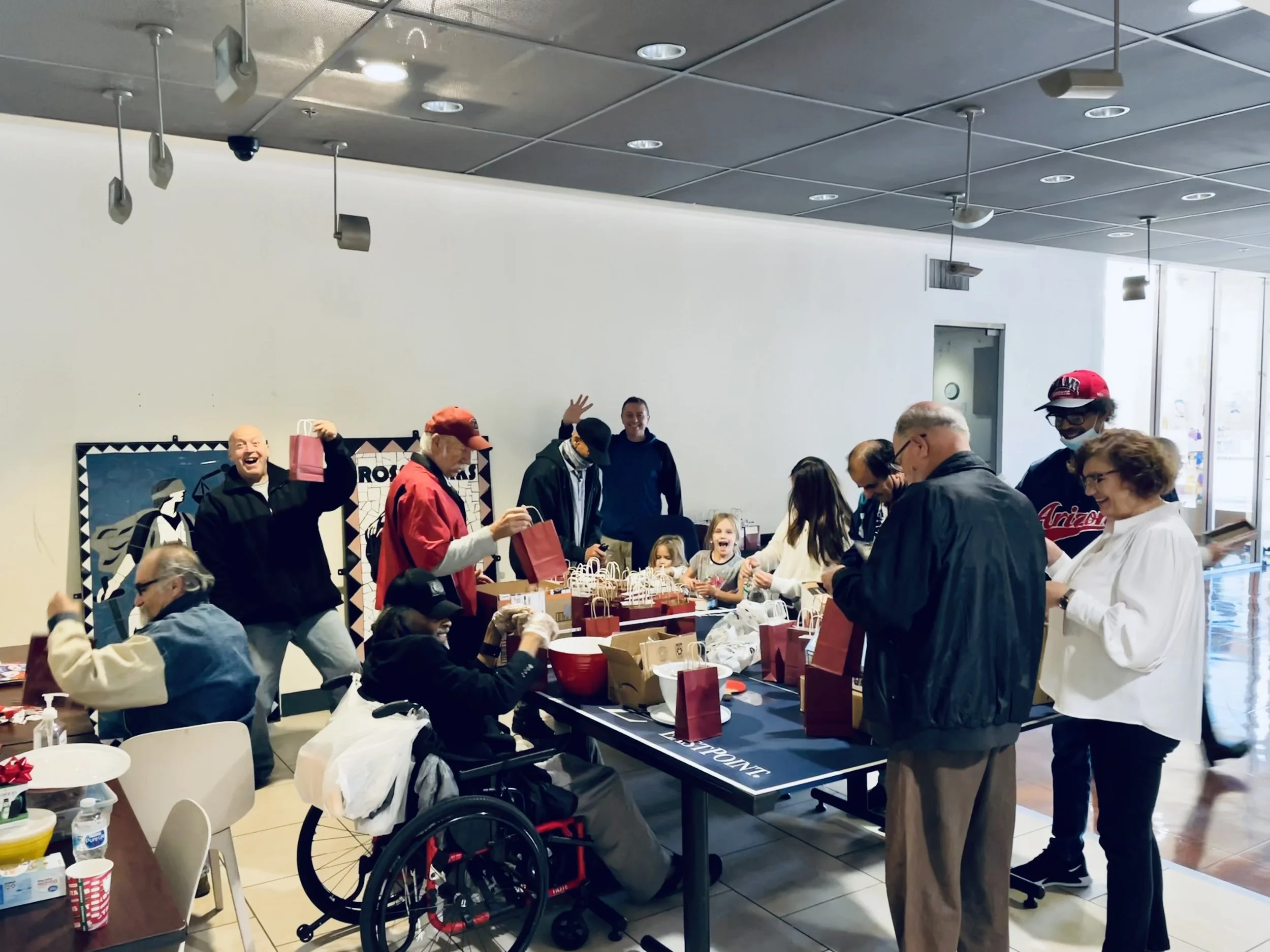 A group of people, including children and adults, are gathered around a table exchanging gifts and celebrating indoors. Some are smiling and there is a festive atmosphere.