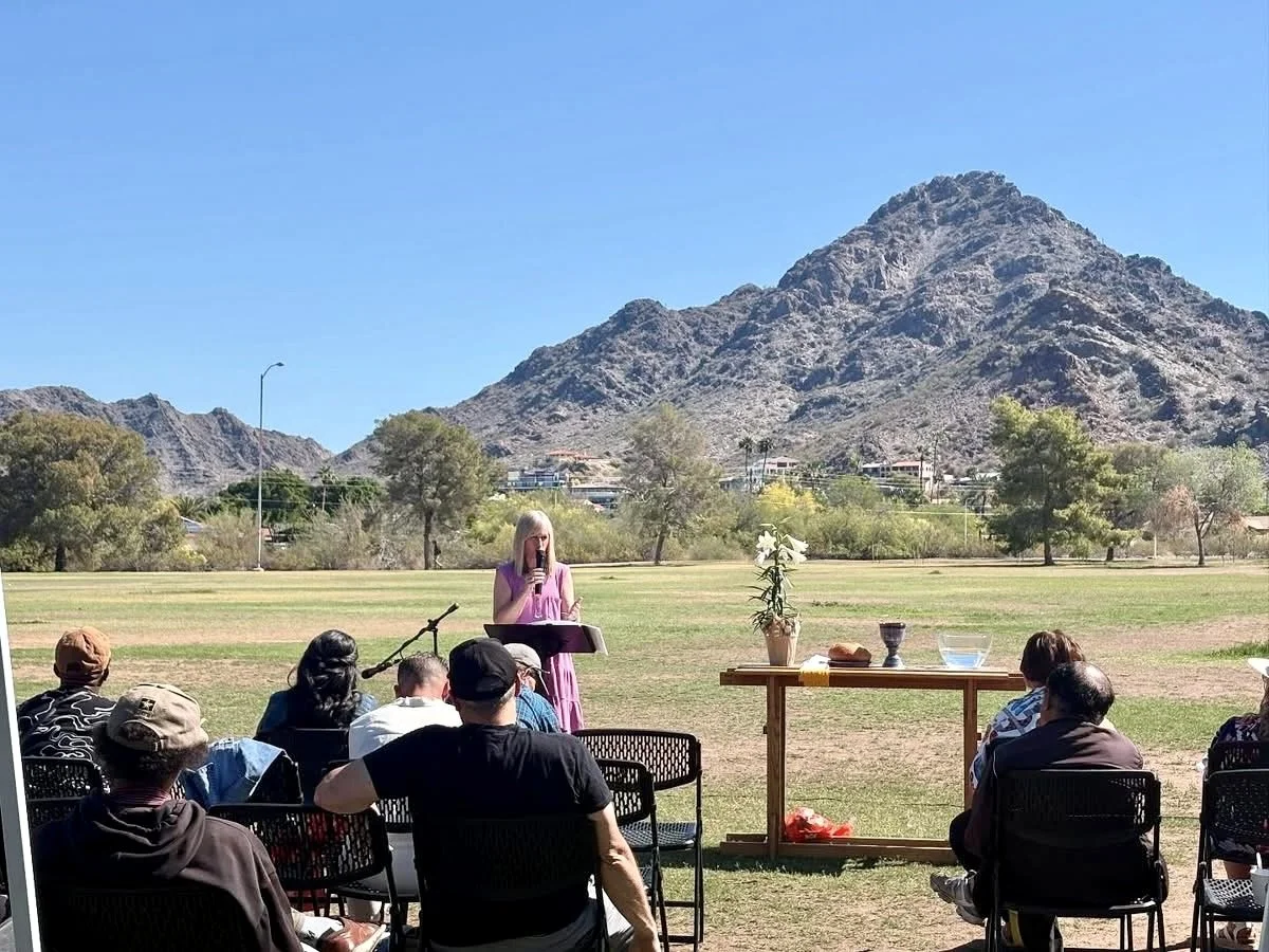 A woman in a pink dress speaking at an outdoor gathering with seated audience in front of her, mountains in the background, and a table with a potted plant and various objects nearby.
