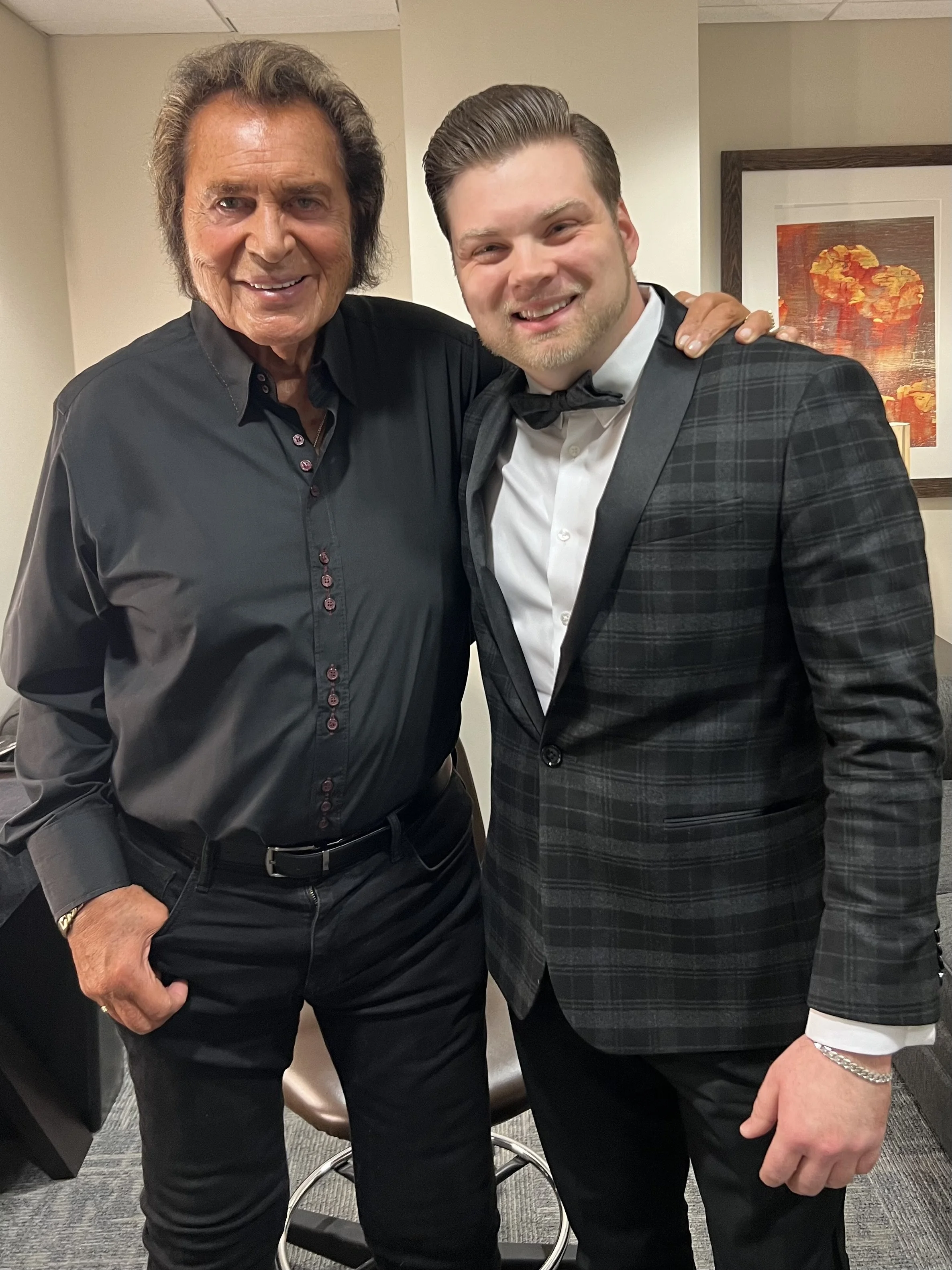 Two men posing together at an event, one older with dark wavy hair in a black button-up shirt, the other younger with light hair in a tuxedo with a bowtie, in an indoor setting with artwork on the wall.