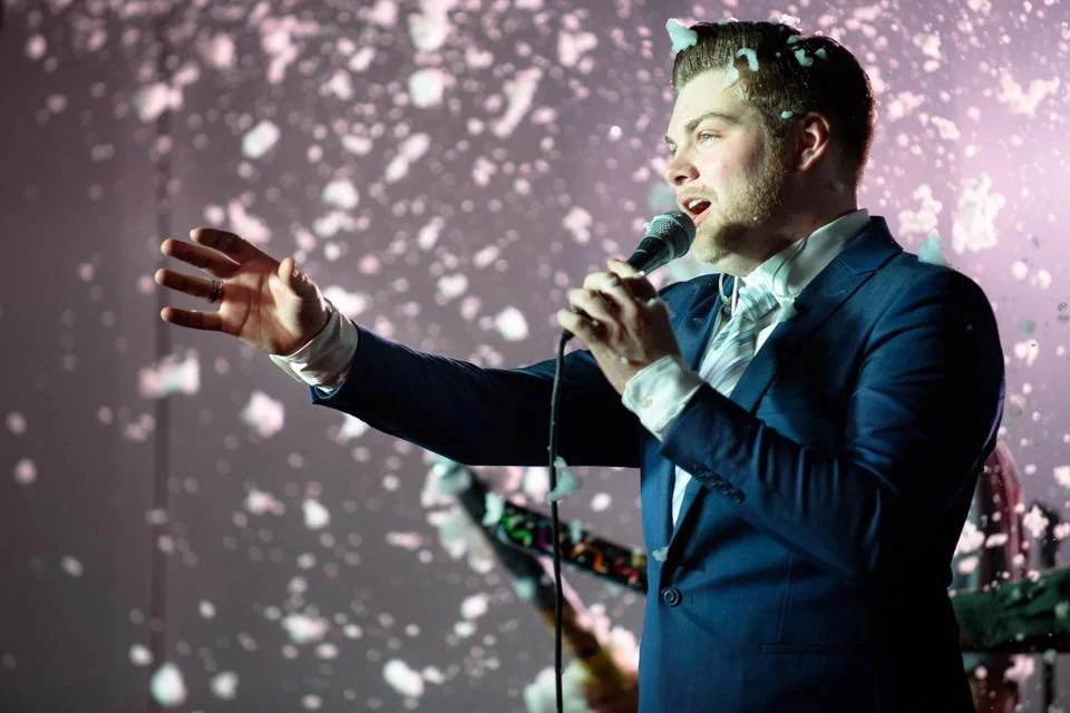 Performer singing into a microphone on stage with a pink and white splattered background.