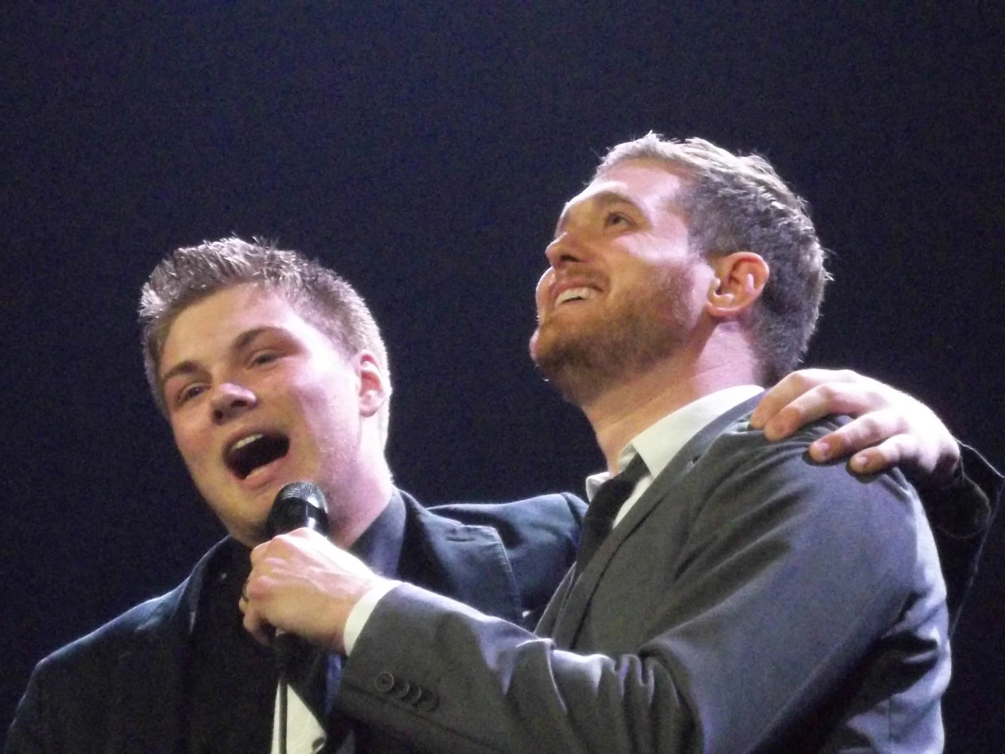 Two men in suits on stage, one holding a microphone, appearing to celebrate or share a moment of joy with the other, who has his arm around him, against a black background.