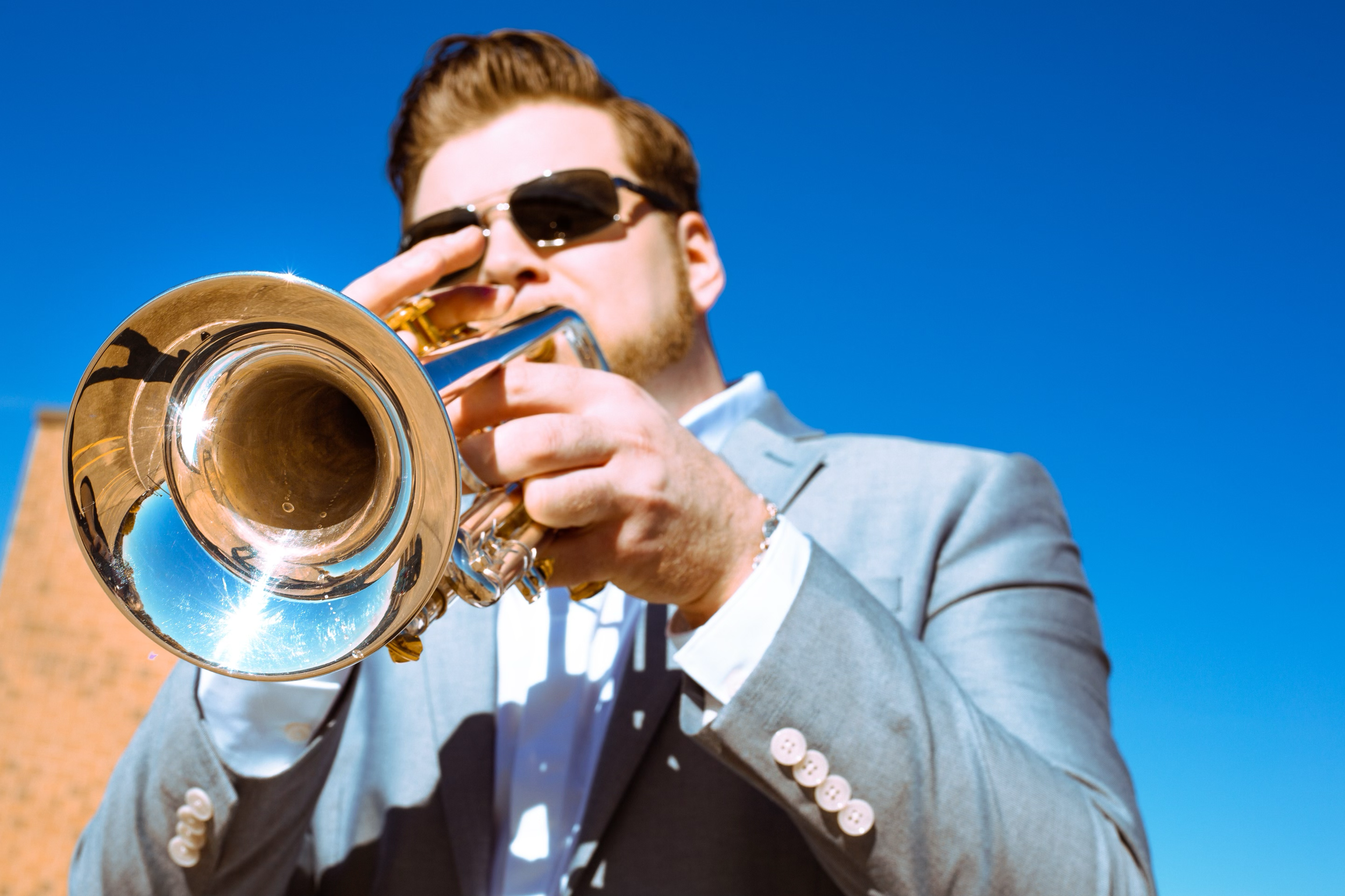 A man in a gray suit and sunglasses playing a trumpet outdoors under a clear blue sky.