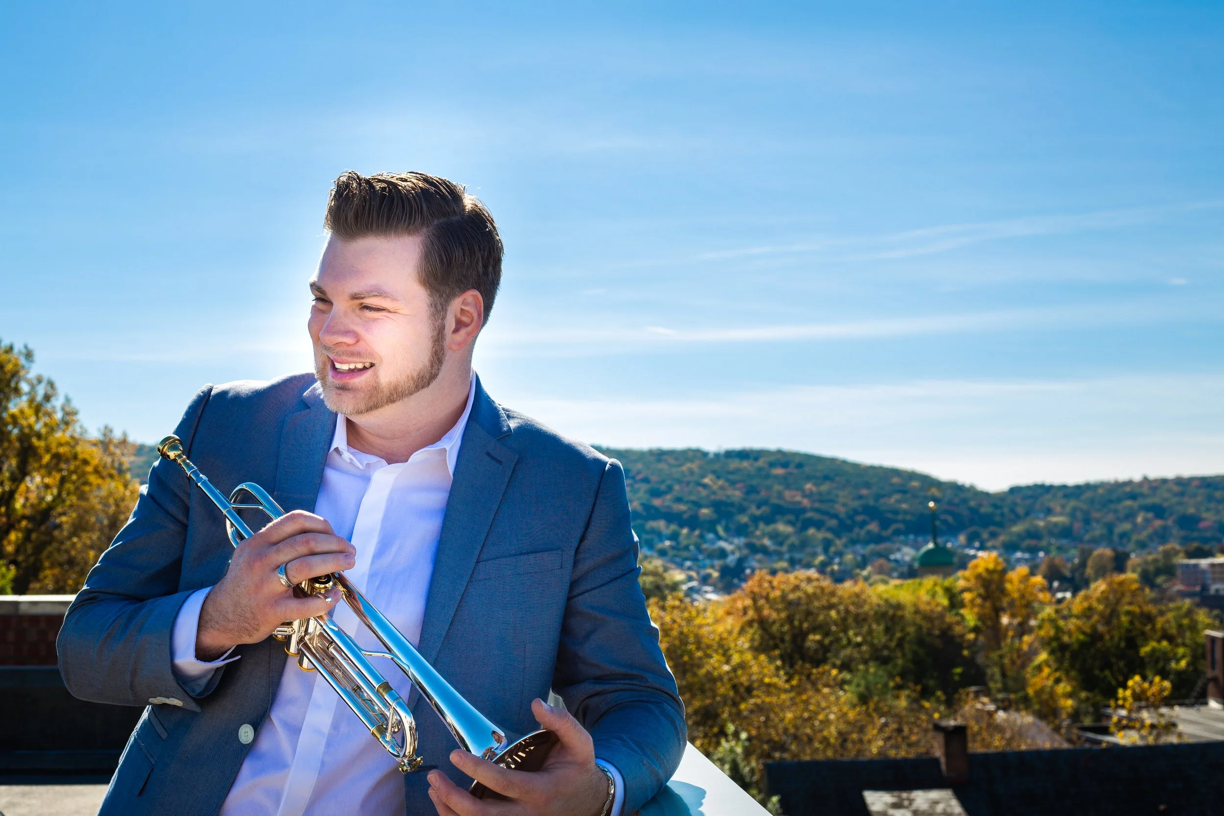 A man in a gray suit holding a trumpet outdoors on a sunny day with trees and hills in the background.