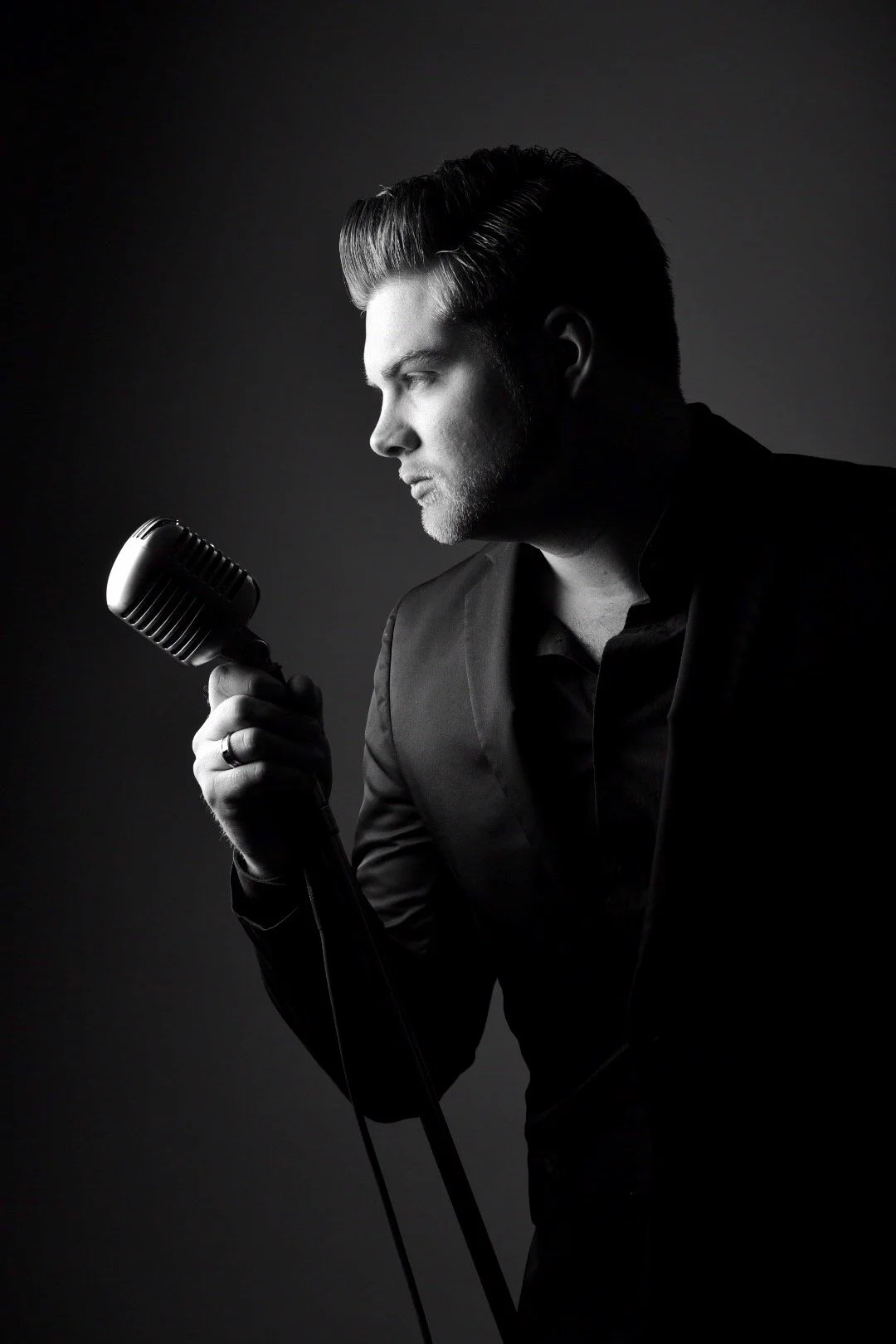 A black and white photo of a man in a suit holding a vintage microphone, looking to the side thoughtfully.
