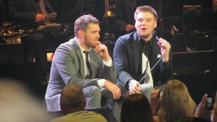 Two men sitting on stage, one holding a microphone, during a live event with an audience in front.