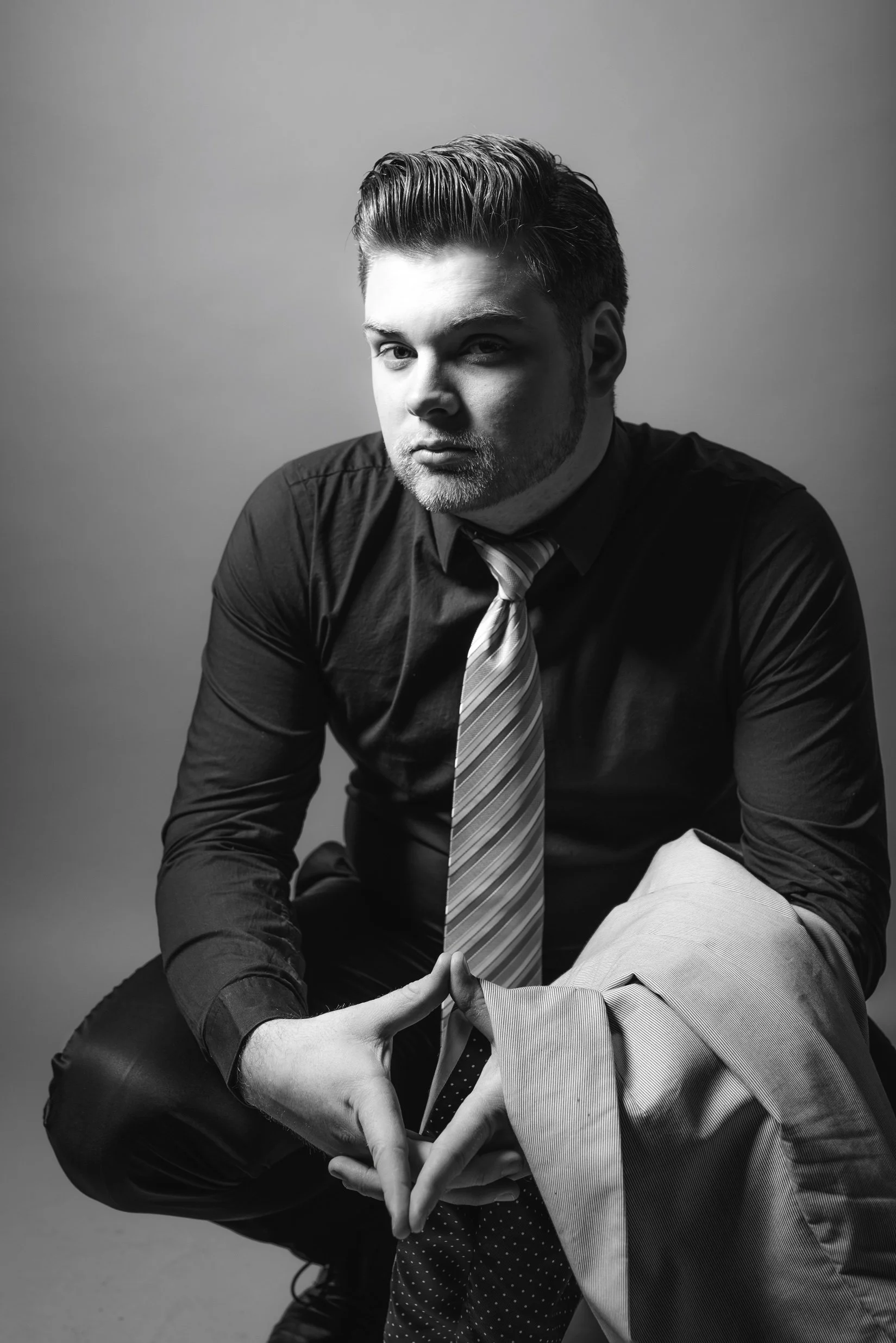 Black and white portrait of a young man with styled hair, wearing a dark shirt and a diagonally striped tie. He kneels with one knee up, holding a light-colored blazer over his arm, and forms a triangular shape with his hands in front of his leg. The