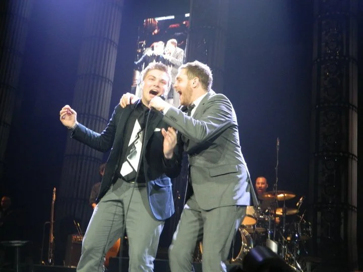 Two men singing together on stage, one in a tuxedo, the other in a suit, smiling and holding a microphone, with a band playing in the background.