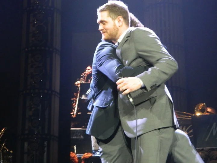 Two men in suits hugging on stage, with a microphone in one man's hand and a jazz band in the background.