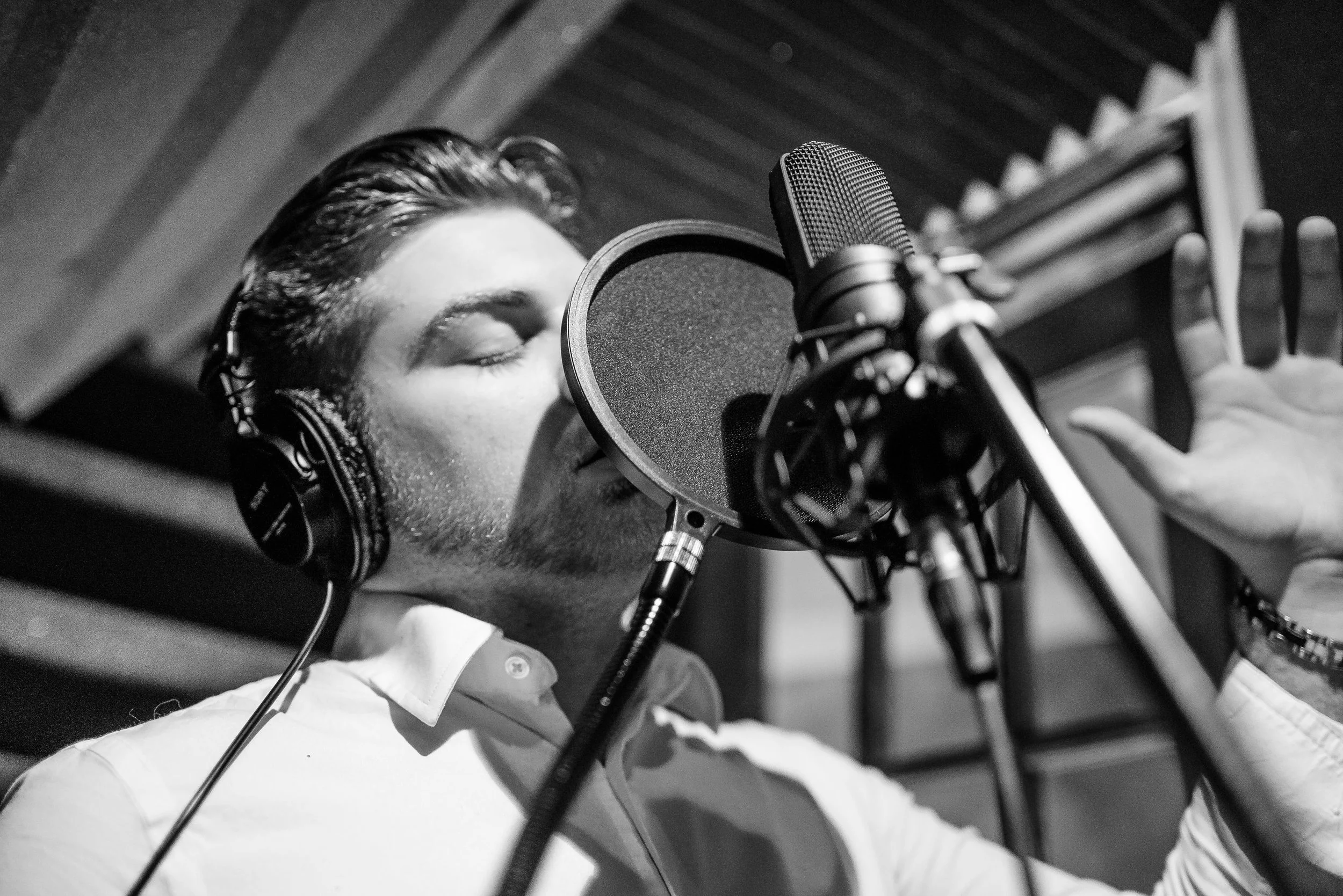 A man in the recording studio singing into a microphone with his eyes closed and wearing headphones.