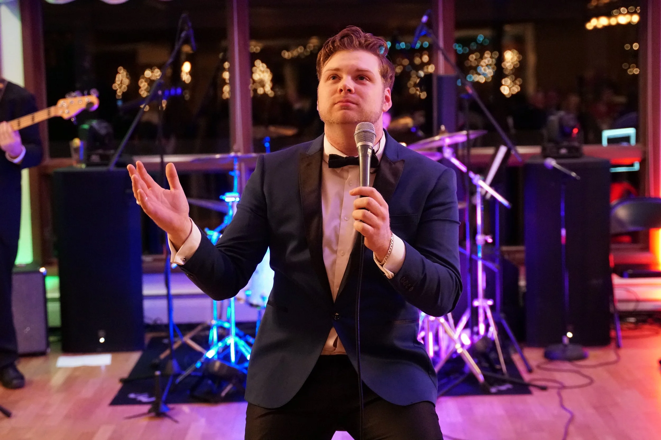 A young man in a tuxedo with a bow tie singing into a microphone at a lively event with band equipment and colorful lighting in the background.