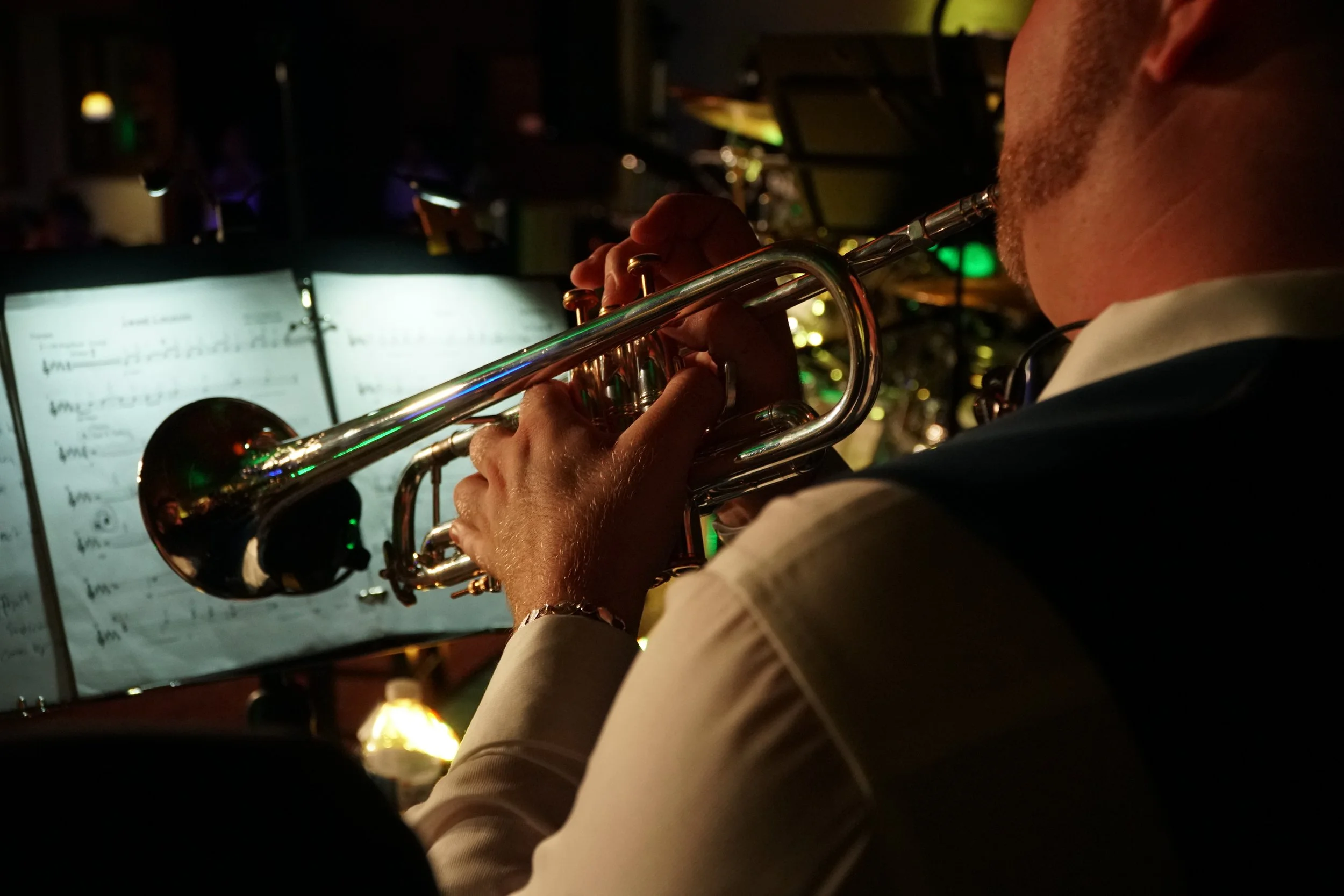 A person playing a trumpet with sheet music in the background, likely in a music venue or during a performance, with dim lighting and stage lights.