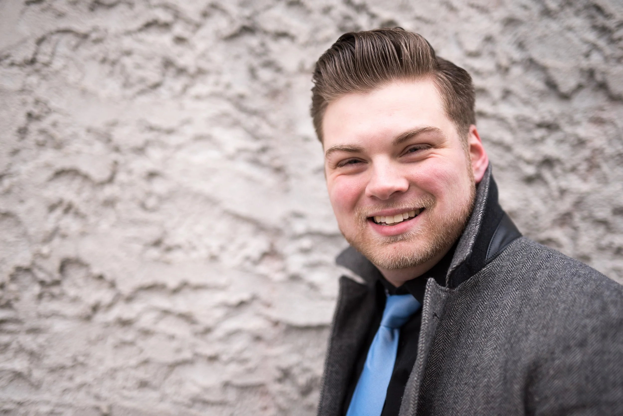 A young man with styled brown hair and a beard, dressed in a gray suit jacket, black shirt, and light blue tie, smiles at the camera against a textured gray stone wall background.