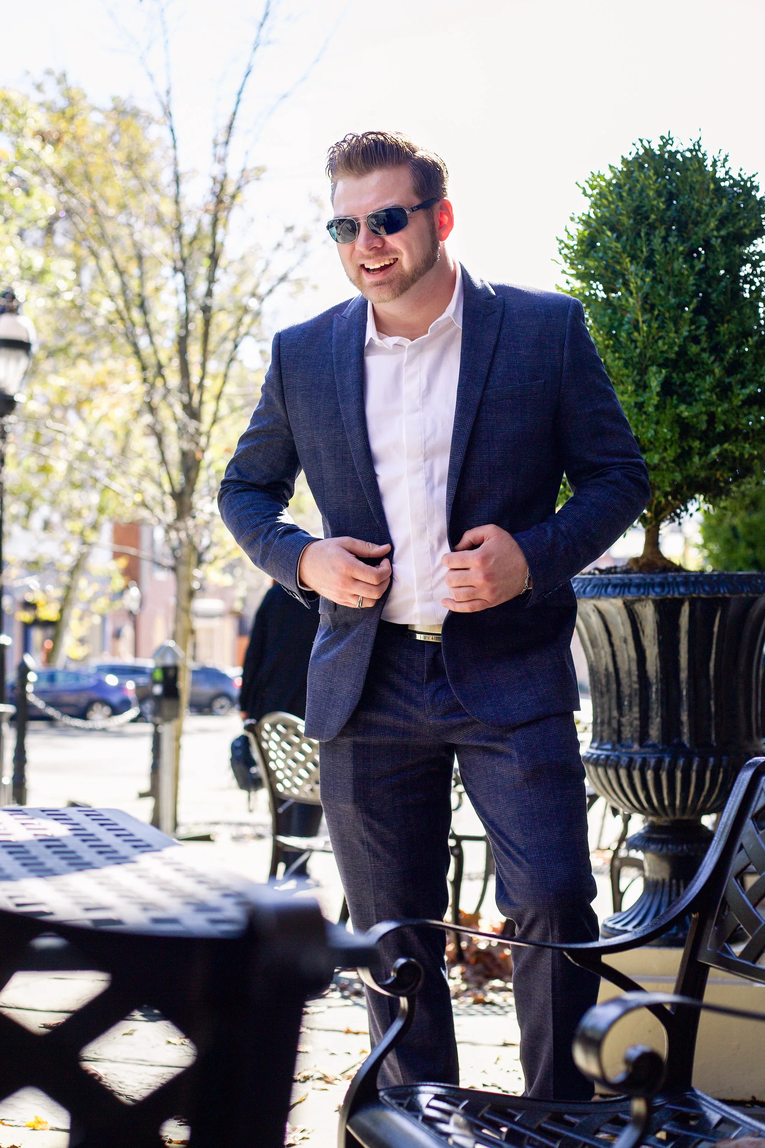 Man in a suit and sunglasses adjusting his jacket outdoors on a sunny day.