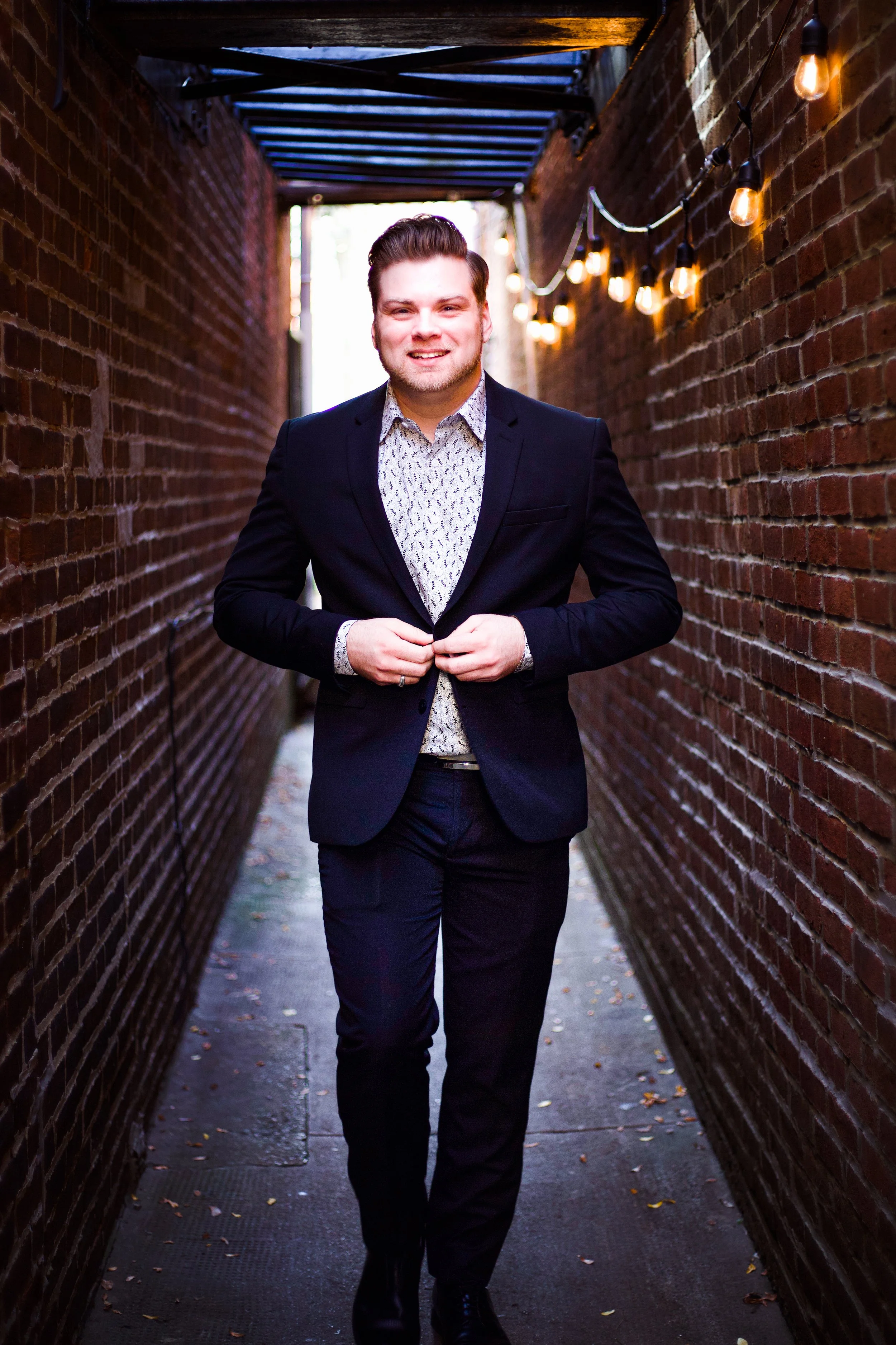 A man in a suit walking through a narrow alleyway with brick walls and hanging string lights.