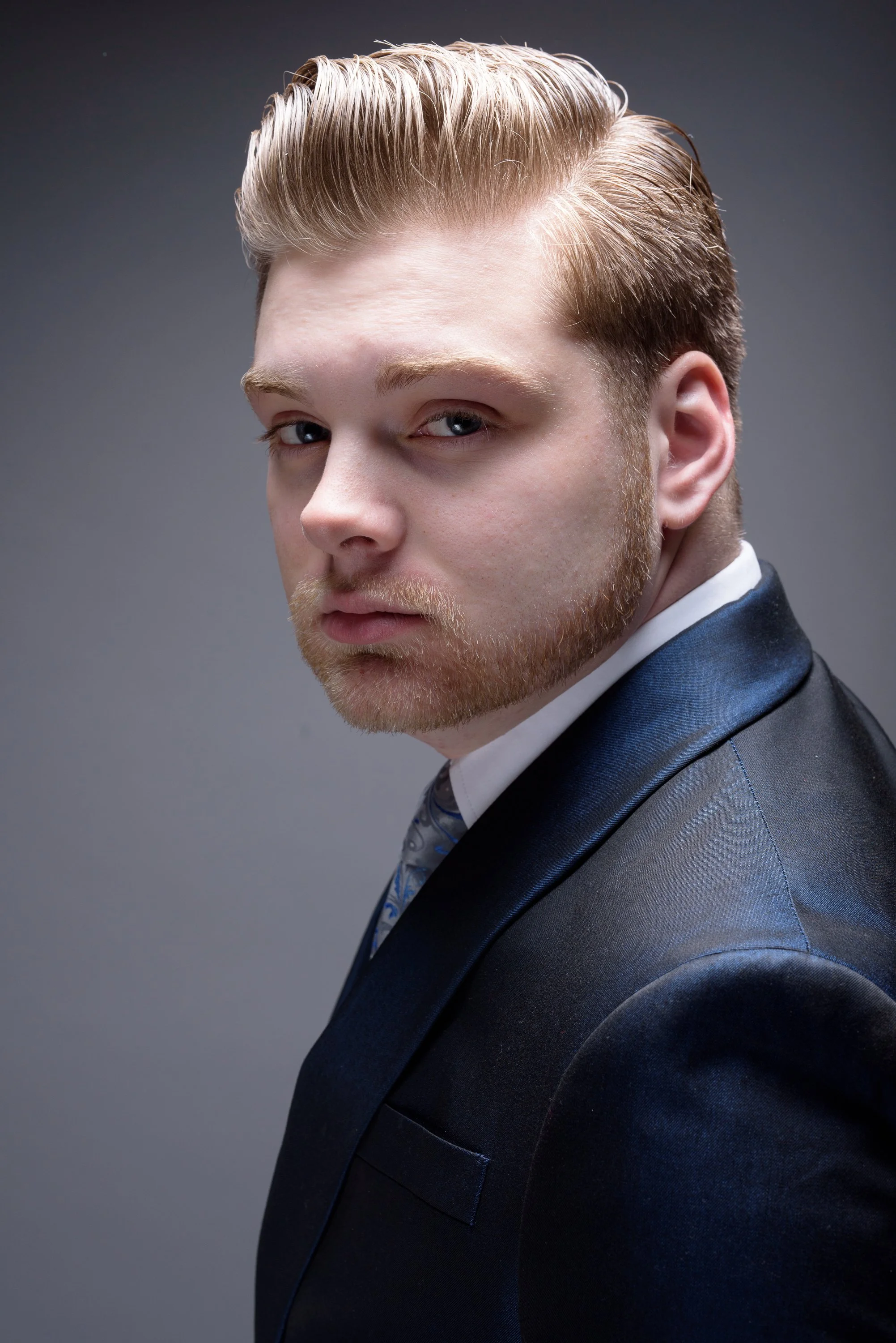 A young man with styled blond hair and a trimmed beard, dressed in a dark suit, white shirt, and patterned tie, posing against a plain gray background.