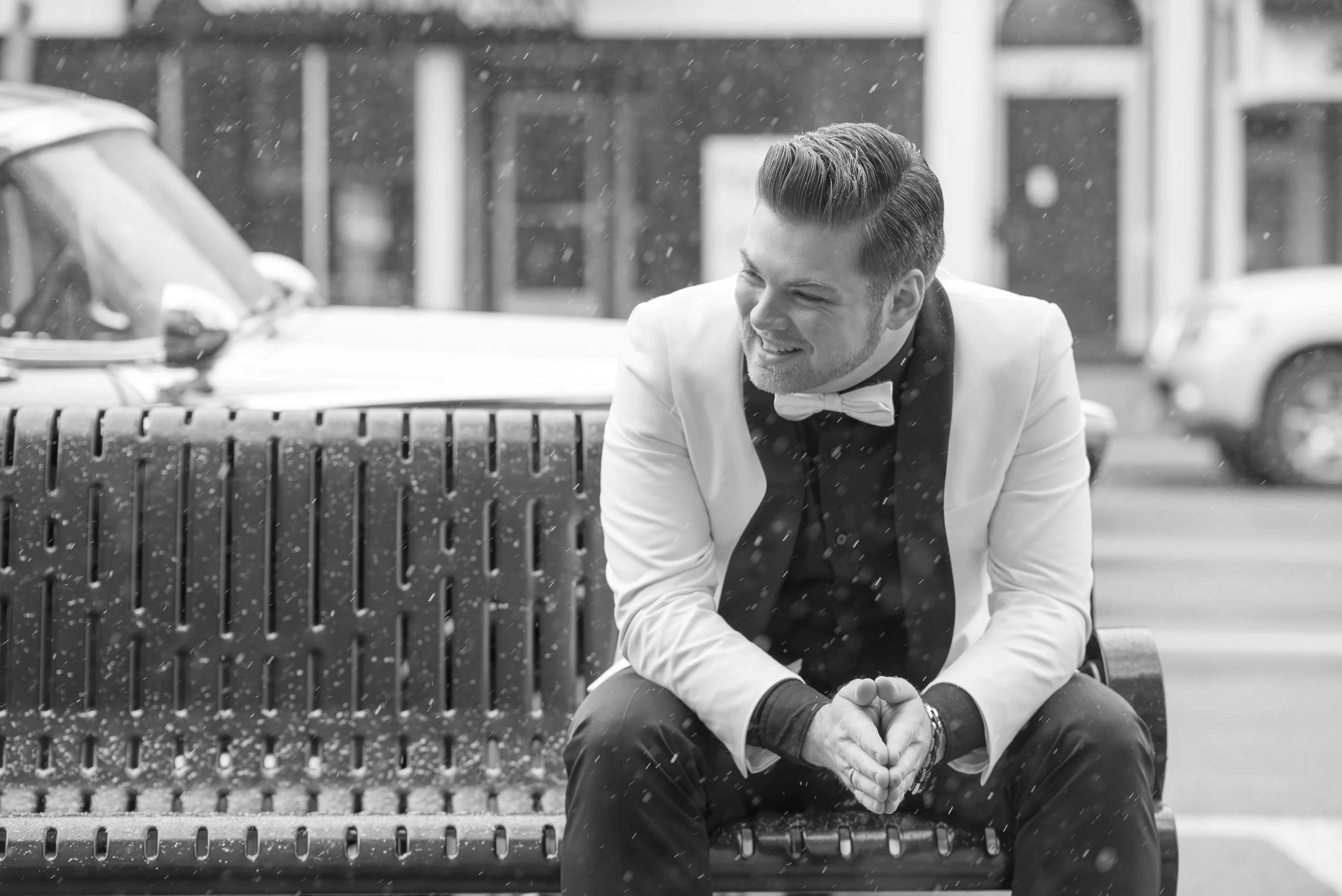 A man dressed in a tuxedo and bow tie sitting on a park bench, smiling as rain falls around him.