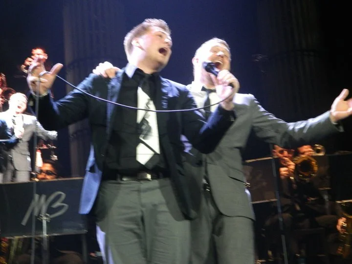 Two male singers in suits perform passionately on stage with microphones, with band members and orchestra in the background.