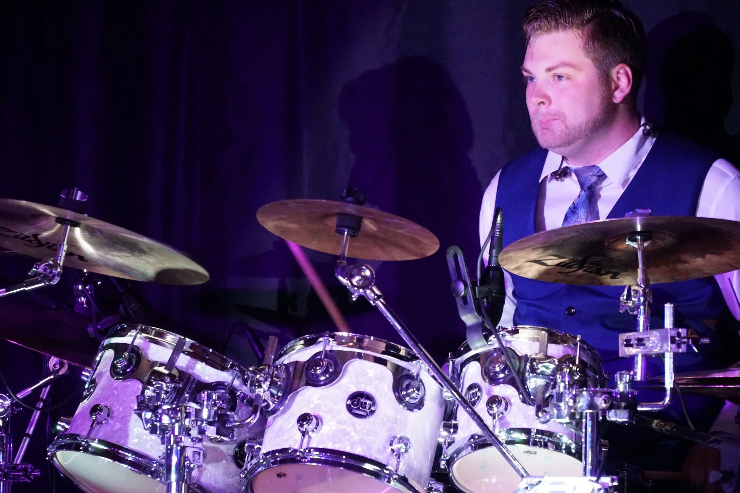 A man in formal attire, including a white shirt, vest, and tie, playing a drum set on stage with dark background and colorful lighting.