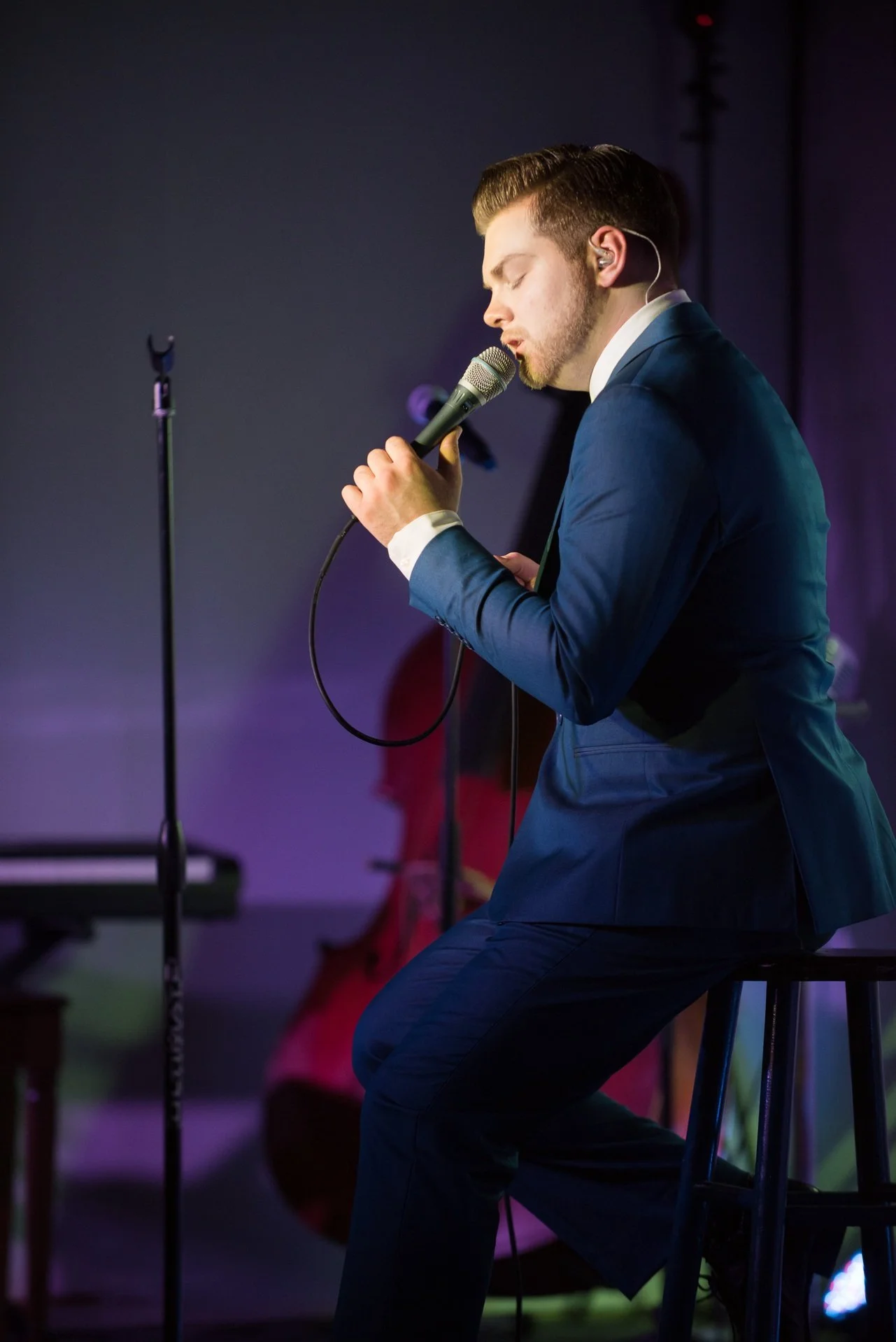 A young man in a blue suit sitting on a stool, singing into a microphone on stage with purple and blue lighting.