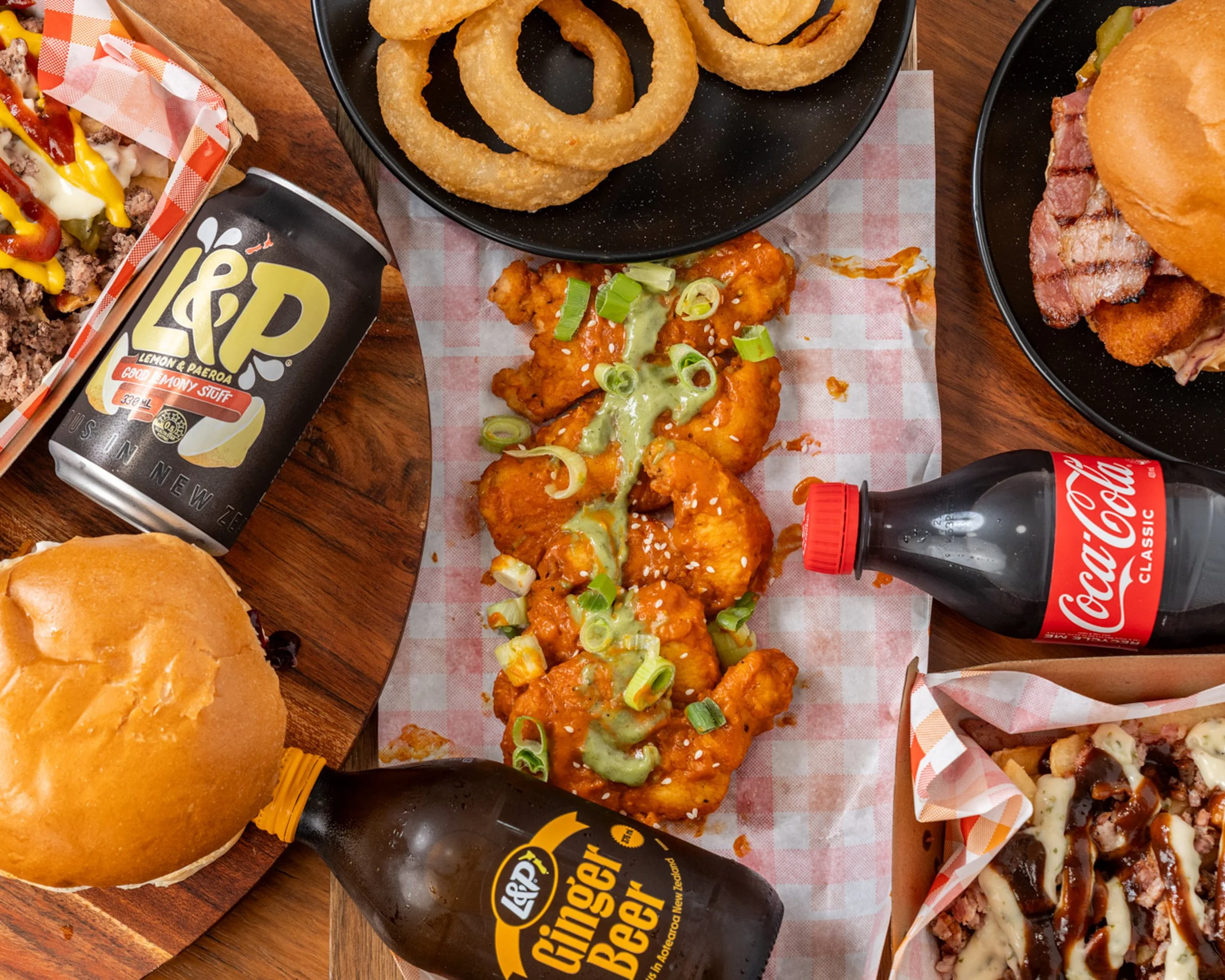 A spread of various foods including onion rings in a black bowl, fried chicken tenders with green onion garnish, cheeseburgers with bacon, and pizza with assorted toppings, along with soft drinks including Coca-Cola, Lemon & Paeroa, and ginger beer.