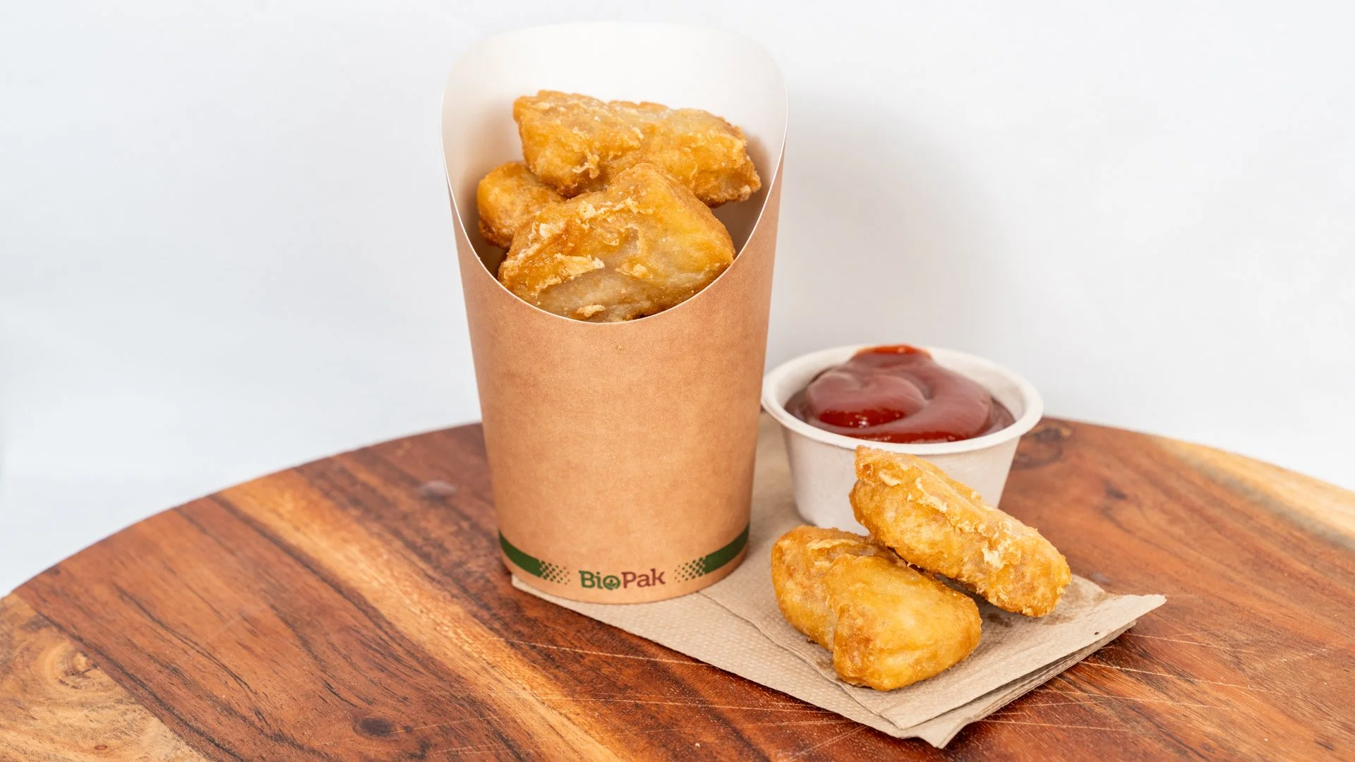 Fried chicken nuggets in a brown paper container, with two nuggets on a napkin, along with a small cup of ketchup on a wooden surface.