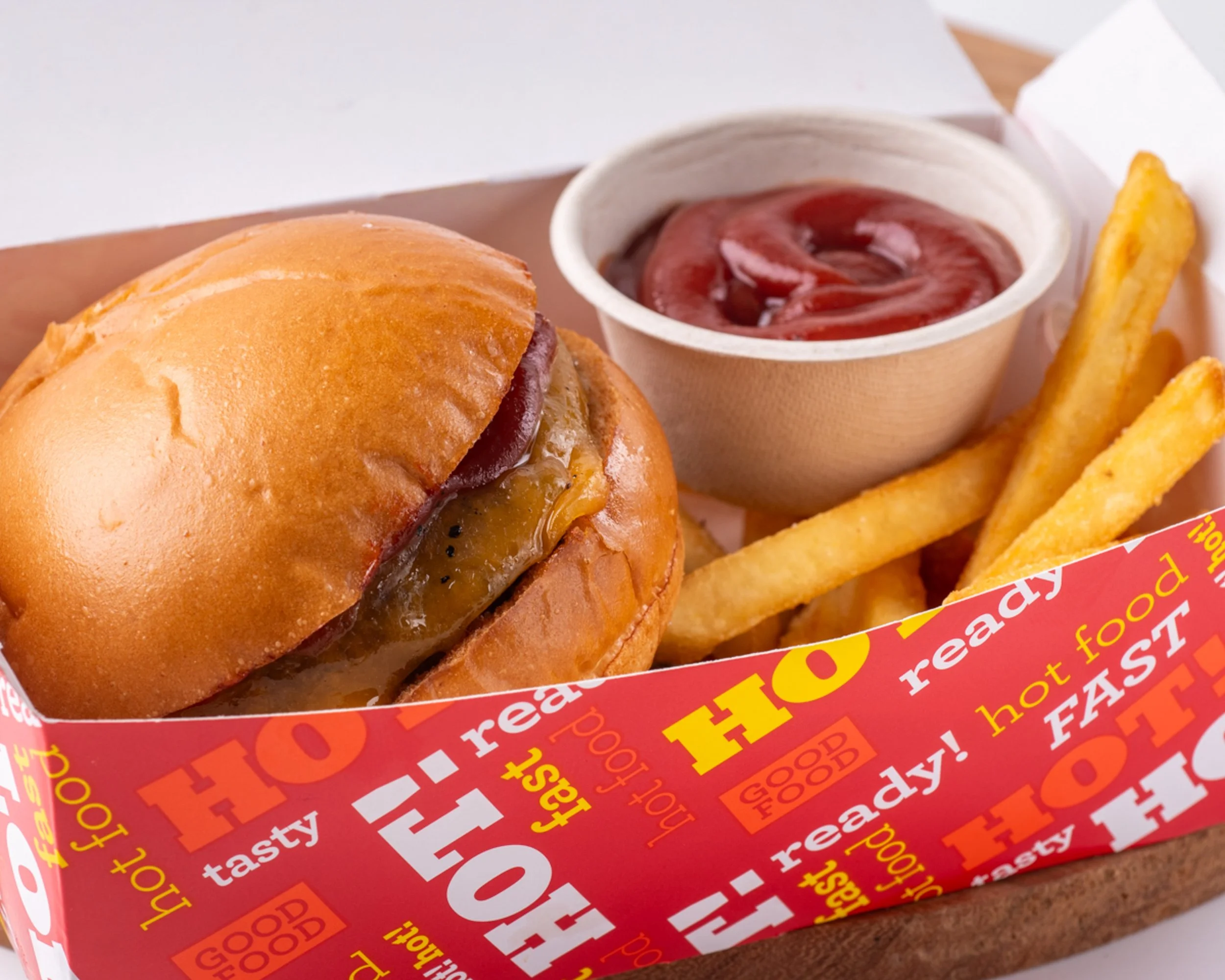 A cheeseburger with barbecue sauce in a soft bun, served with French fries and ketchup in a small paper bowl.