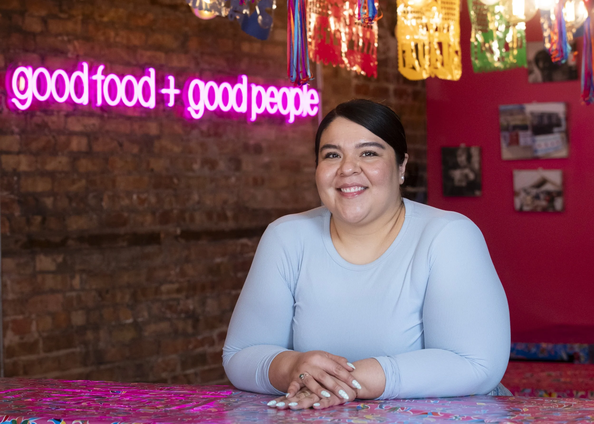 A smiling woman with dark hair sitting at a colorful table in front of a brick wall with a pink neon sign that reads 'good food + good people.' Cheerful decorations hang from the ceiling in a festive setting.