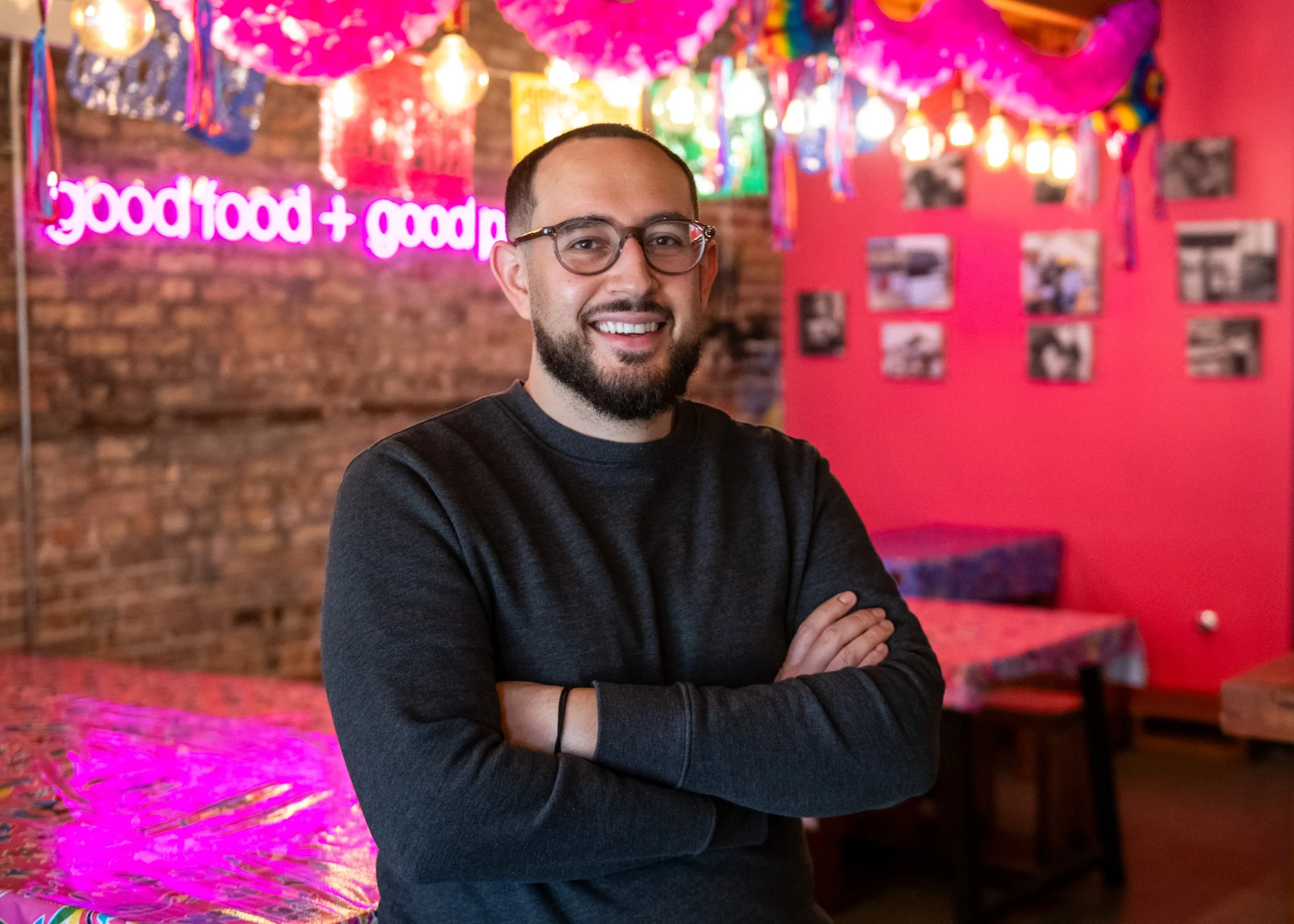 A man with glasses and a beard wearing a black long-sleeve shirt, smiling with arms crossed, standing indoors in front of colorful decorations, including pink, purple, and green lighting, a pink wall with framed pictures, and a sign that says 'good food' in neon lights.