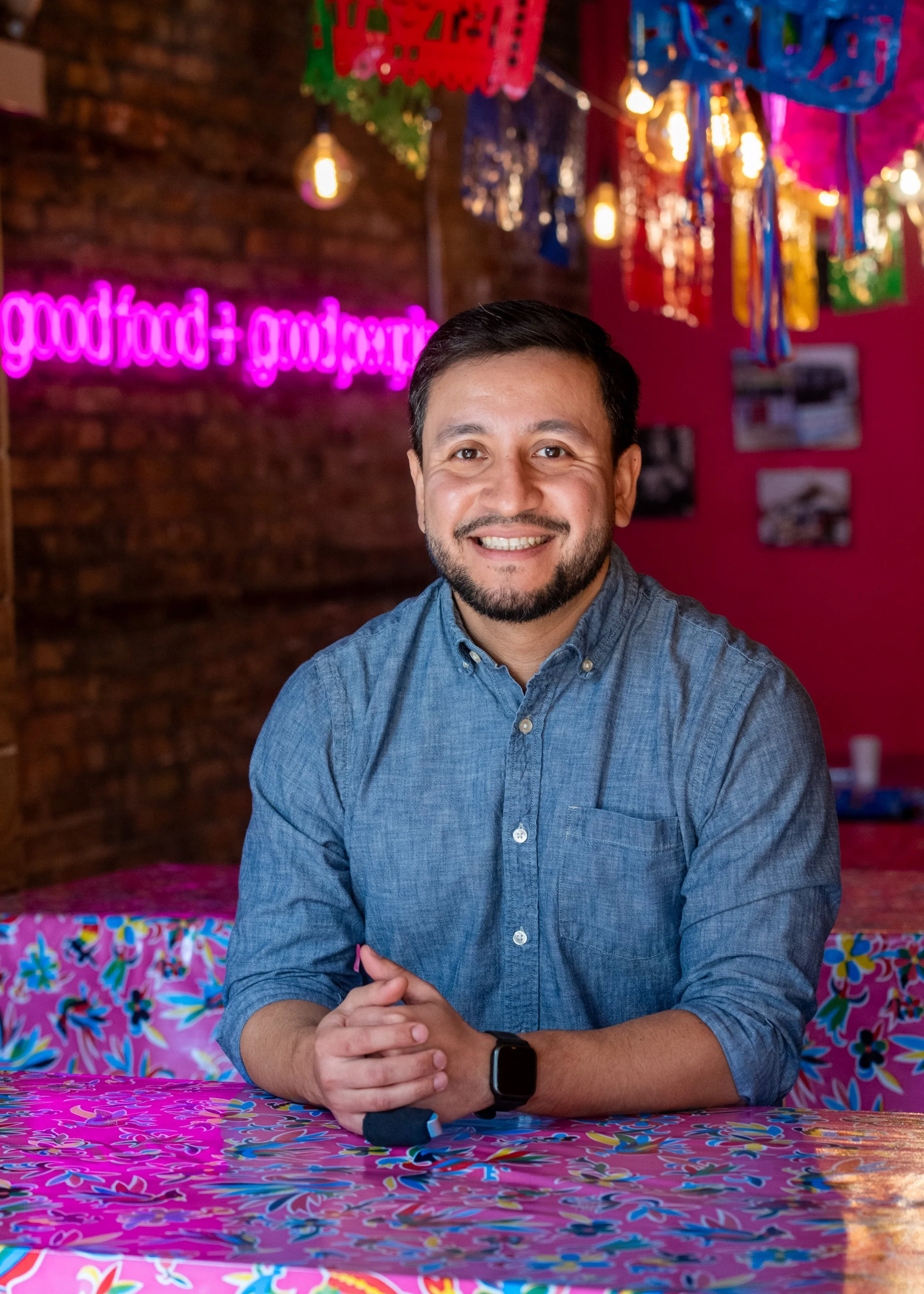 A smiling man with dark hair and a beard, wearing a blue denim shirt and a smartwatch, sitting at a colorful table with a vibrant background of pink, red, and green decorations, and a glowing pink sign that reads 'food + drink'.