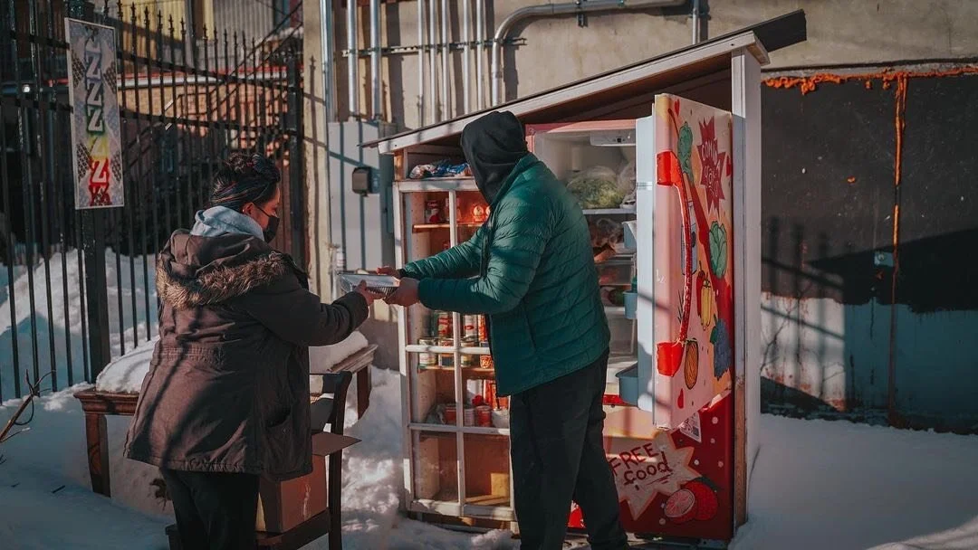Person in a green jacket receiving a food item from another person in a winter coat outside a small open refrigerator with bright colors and graphics, in snowy conditions.