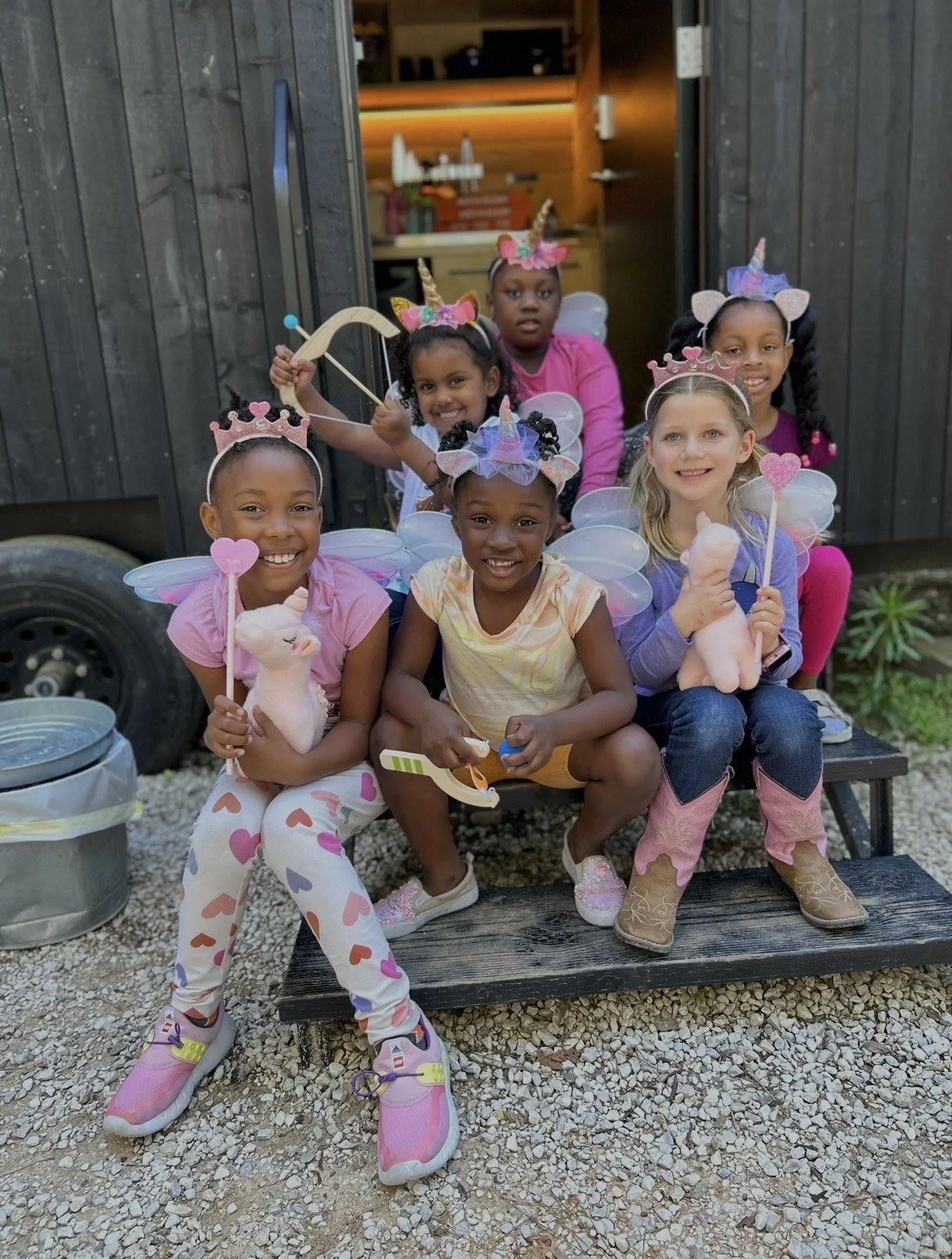 Six children dressed as unicorns with wings and fairy crowns, holding plush unicorn toys and wands, sitting on a bench in front of a wooden shed during a birthday party.